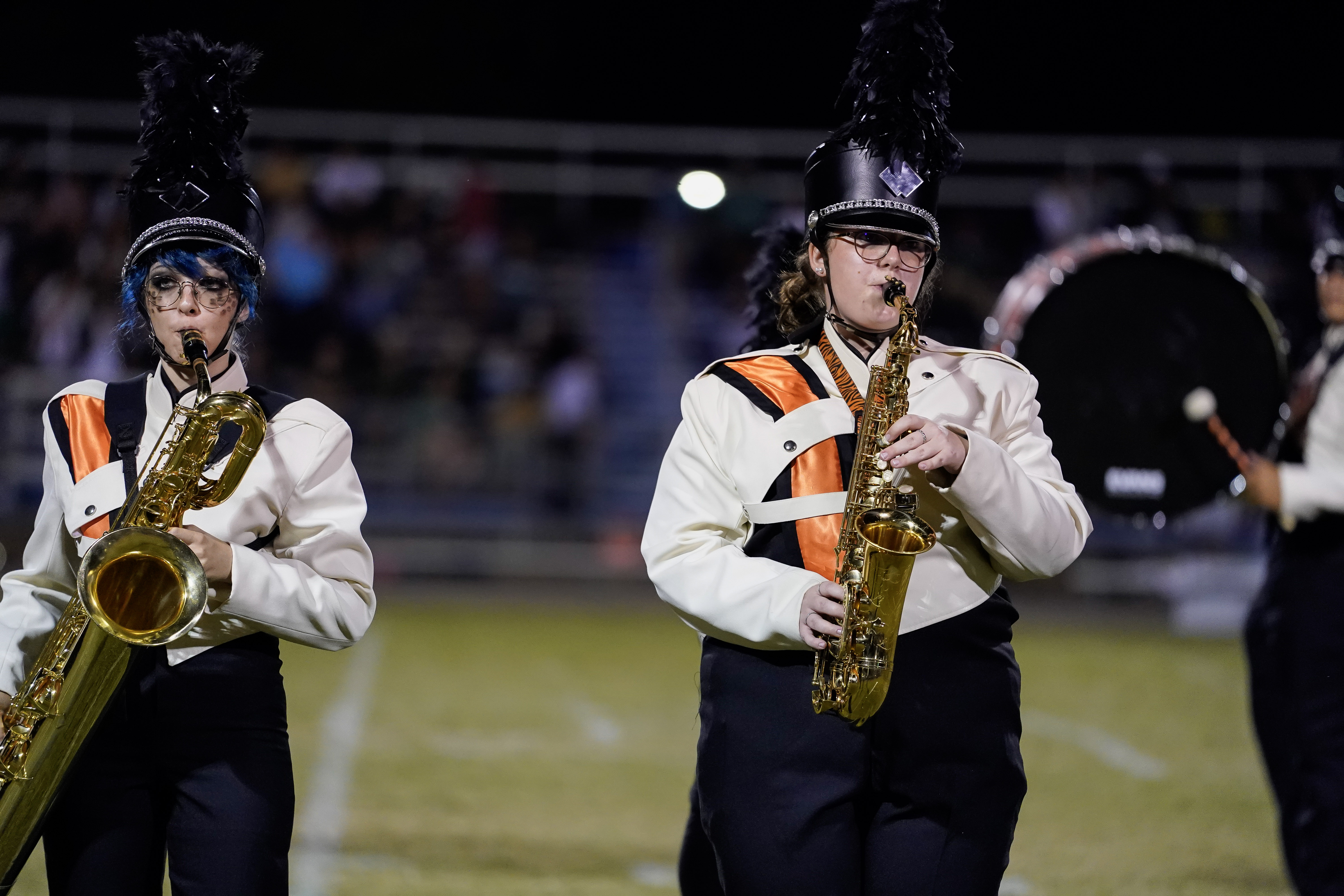 Two marching band members in black hats with plumes and cream uniforms with orange and black sashes play saxophones on a field at night. A blurred drum and audience are visible in the background.