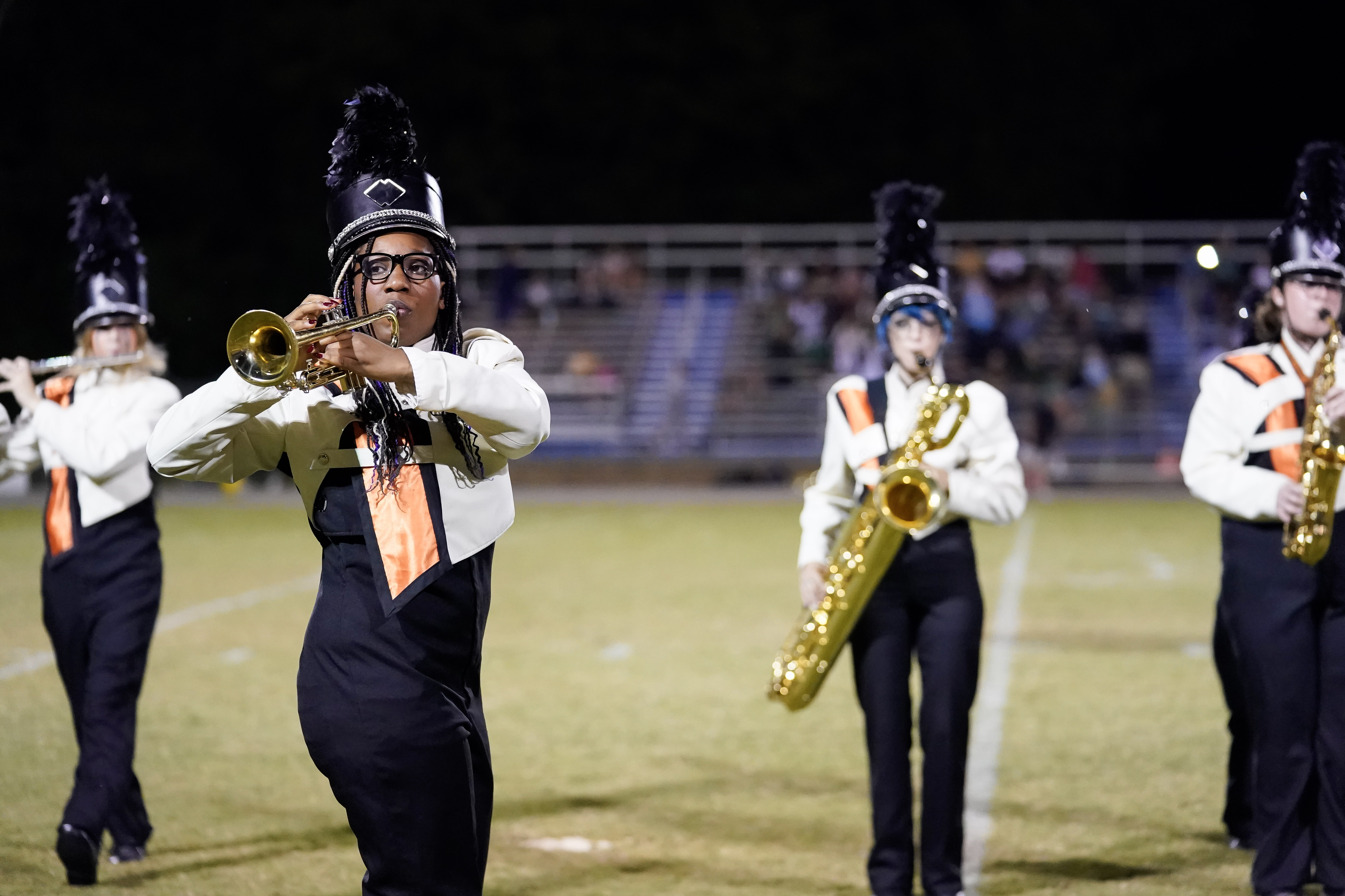 A marching band performs on a football field at night. A trumpet player is in the foreground, with other band members playing saxophones in the background. All are wearing uniforms and feathered hats.