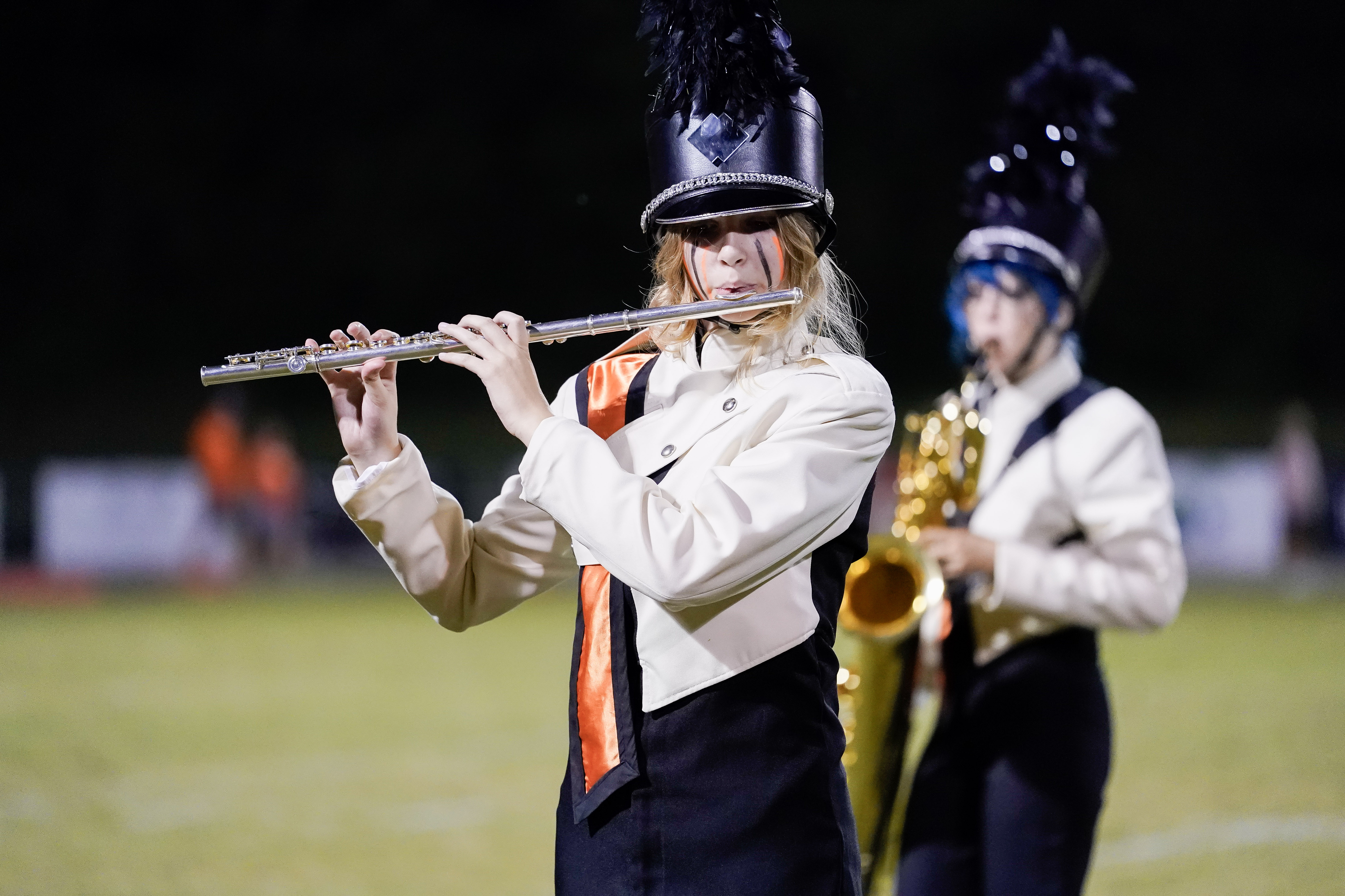 A marching band member in uniform plays the flute on a field, with another band member playing saxophone in the background.