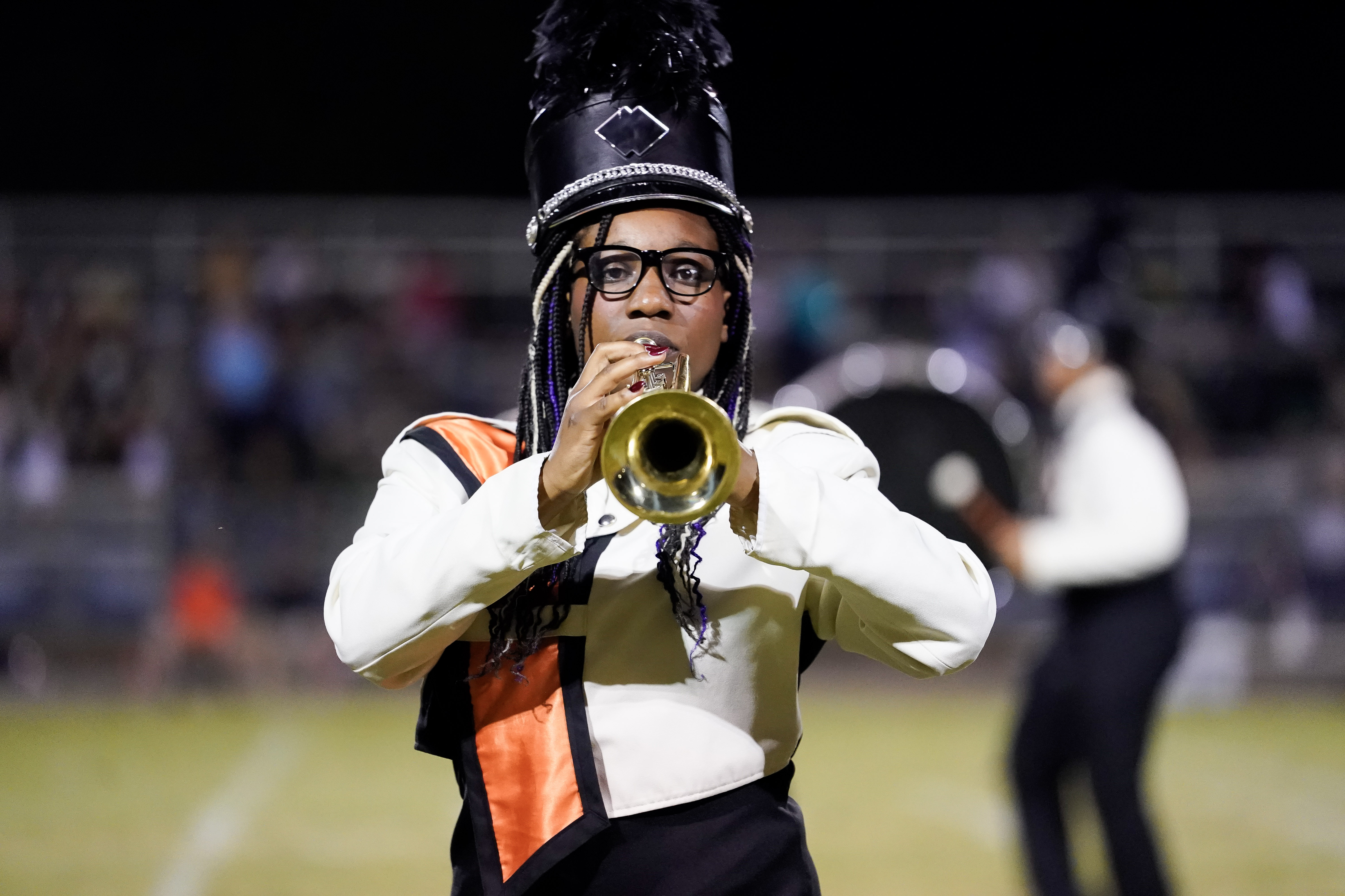 A marching band member in a black and white uniform with an orange sash plays a trumpet on a field at night. The band member wears glasses and a tall hat with a plume. Another band member is blurred in the background.