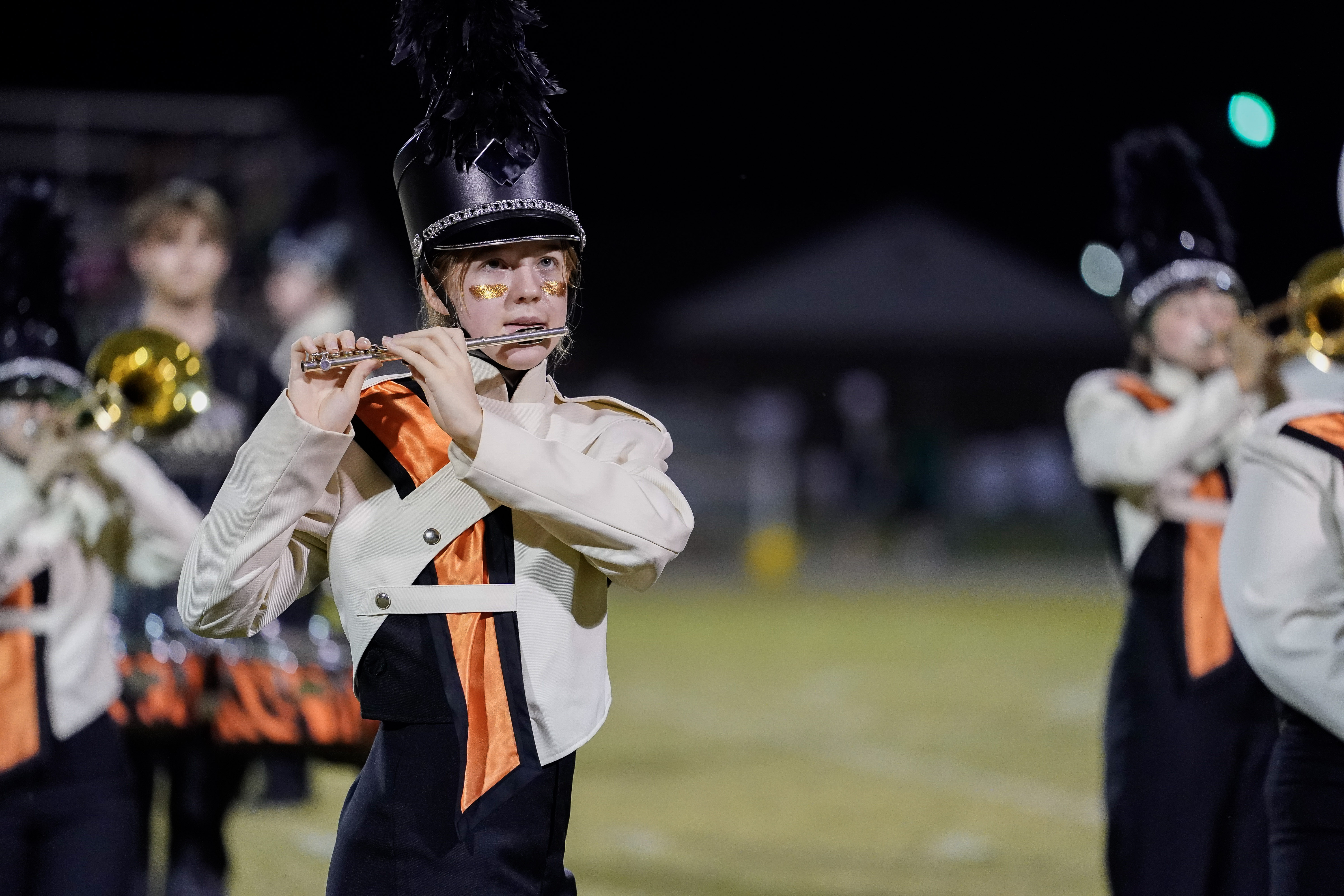 A marching band member in a black and white uniform with orange accents plays a flute on a field at night. Other band members are visible in the background, also playing instruments.