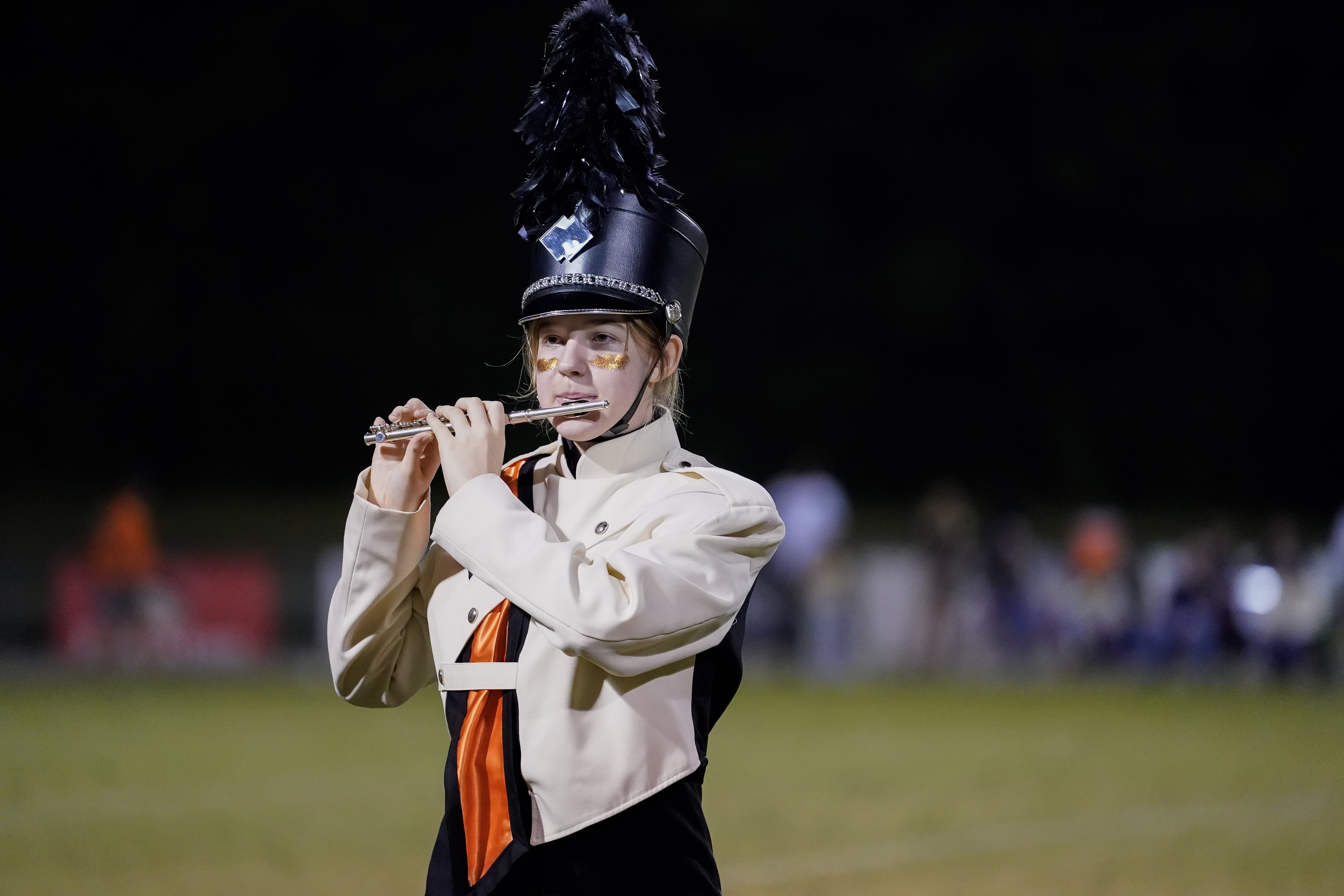 A marching band member in a cream and black uniform with a tall hat plays a flute outdoors at night, standing on a grassy field with blurred people and trees in the background.