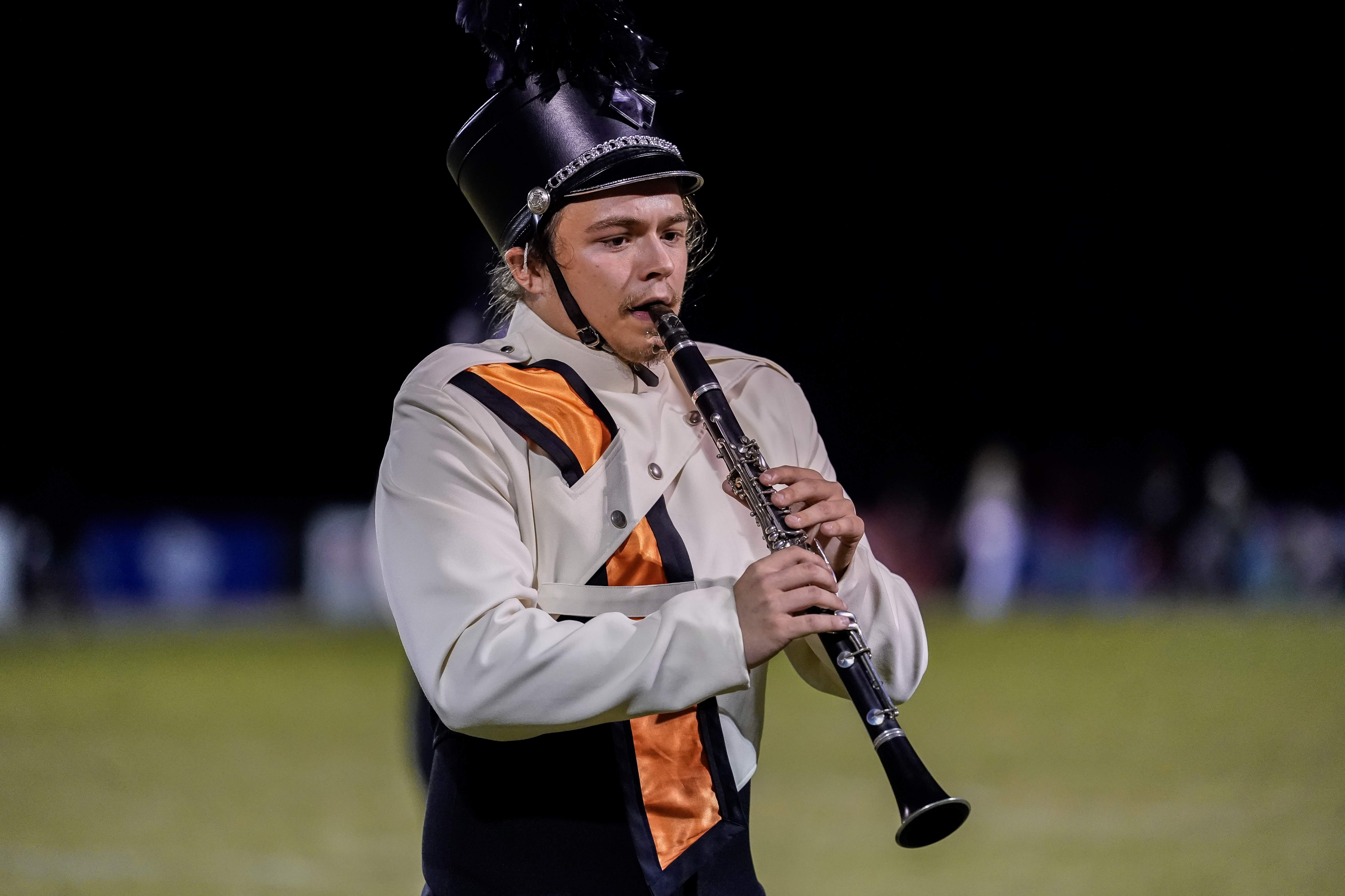 A marching band member in a white and black uniform with orange accents plays a clarinet on a field at night.
