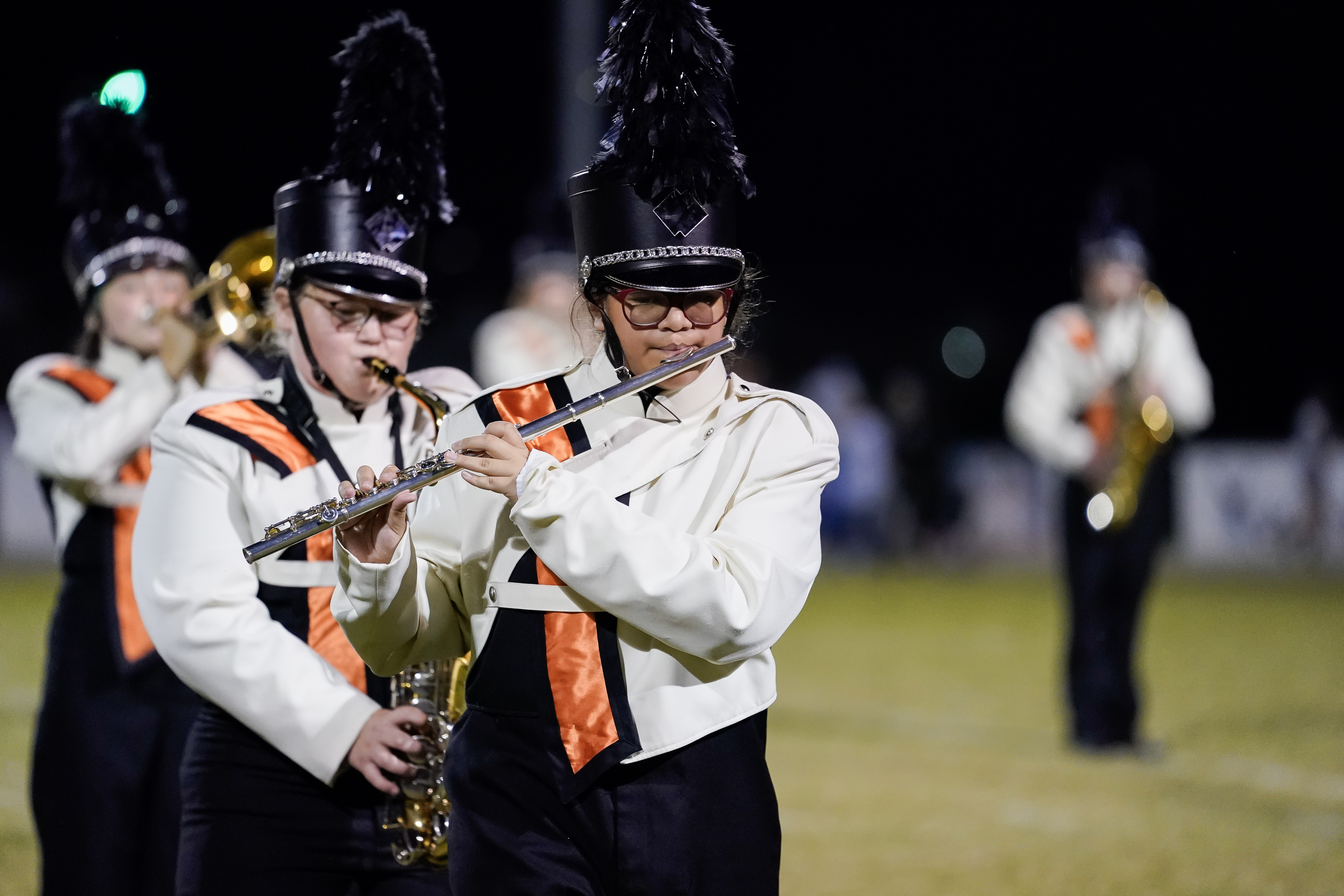 A marching band performs at night on a field. The focus is on a flute player in a white uniform with black and orange accents, wearing a tall black plume hat, with other band members playing behind them.