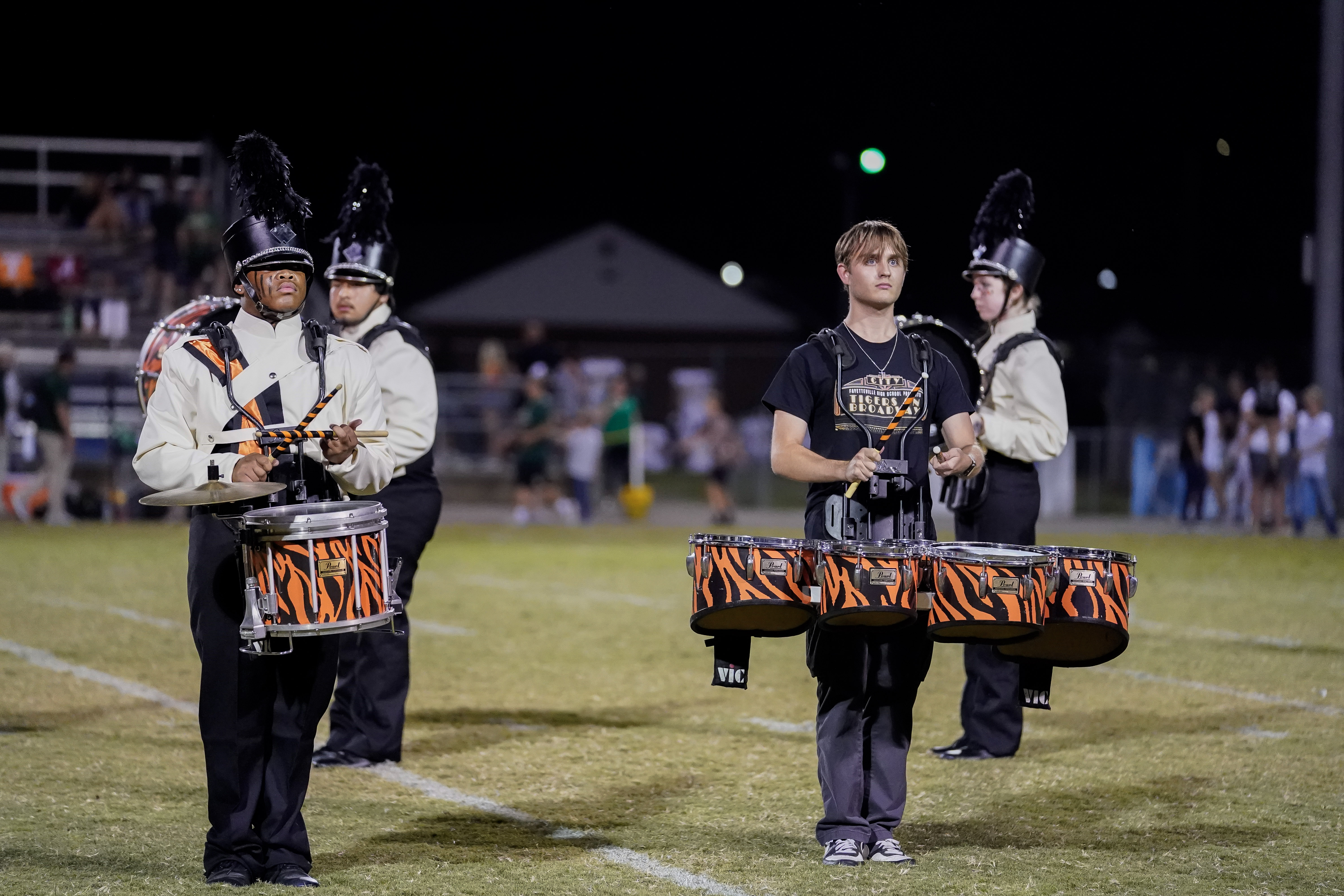 A marching band performs on a field at night. A drummer with tiger-striped drums stands in the center, surrounded by other band members playing instruments in black and orange uniforms.