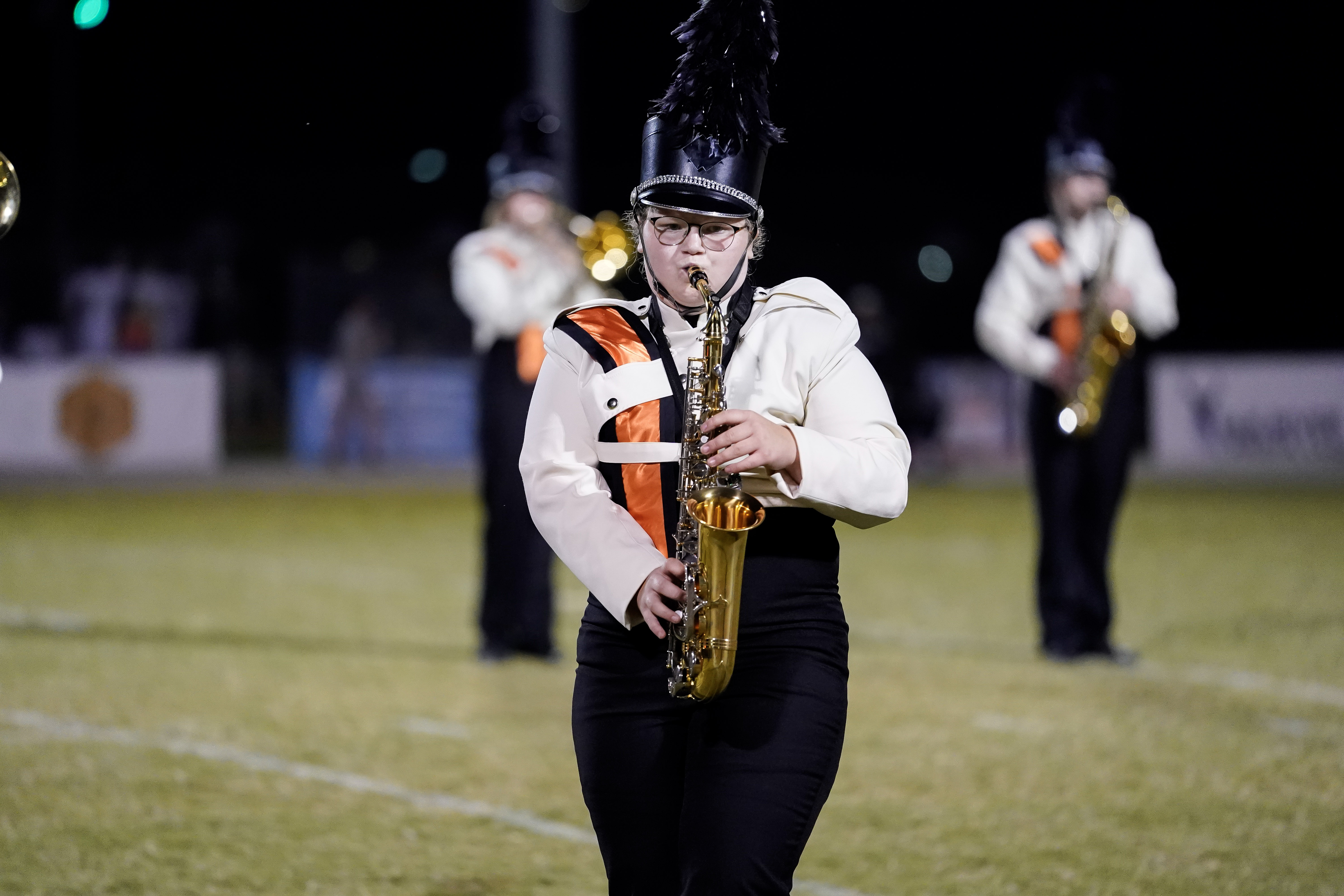 A marching band member in uniform plays a saxophone on a football field at night, with other band members visible blurred in the background.