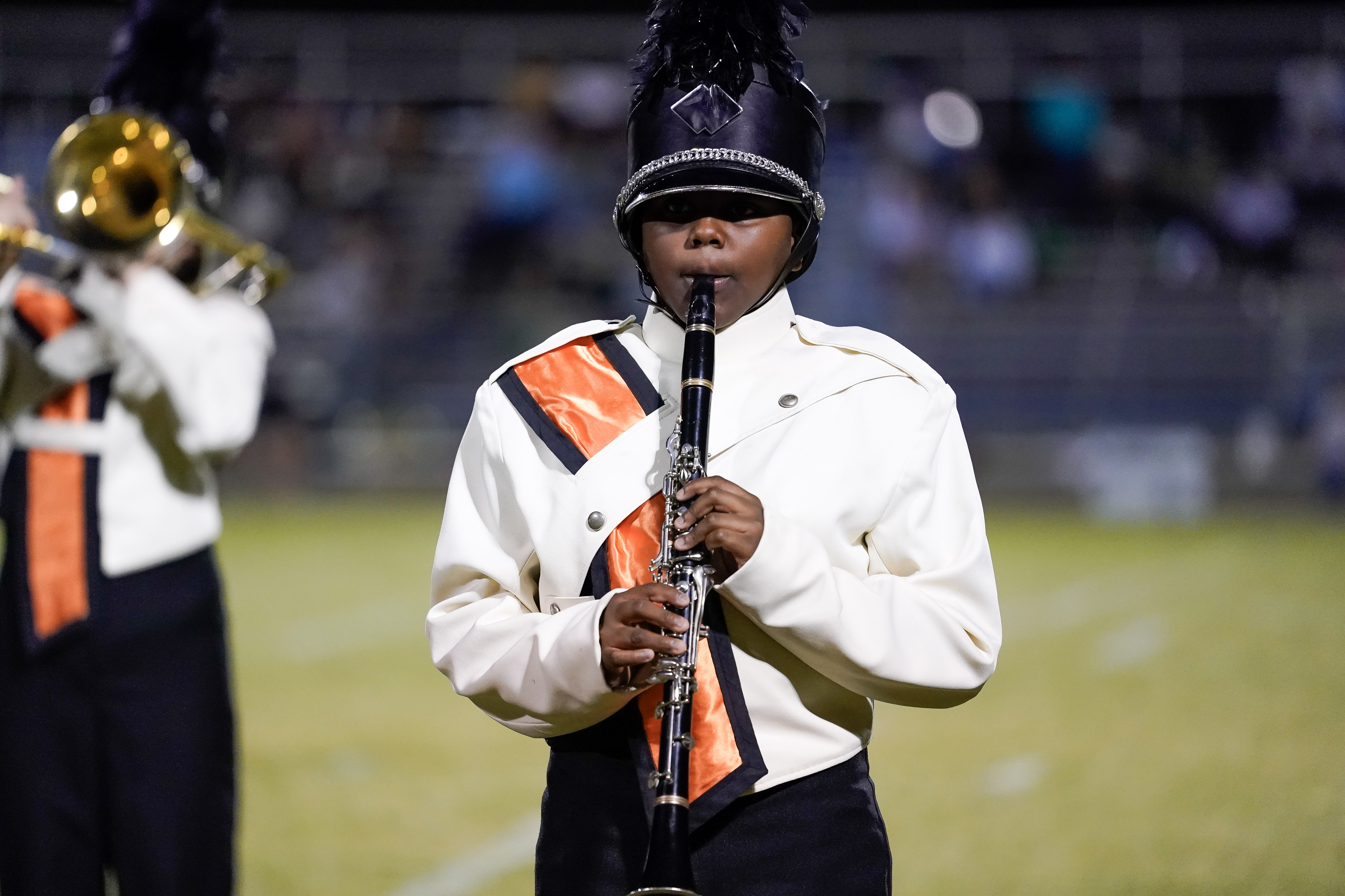 A marching band member in a white and black uniform with orange accents plays a clarinet on a field during a performance. Another band member holding a brass instrument is partially visible in the background.