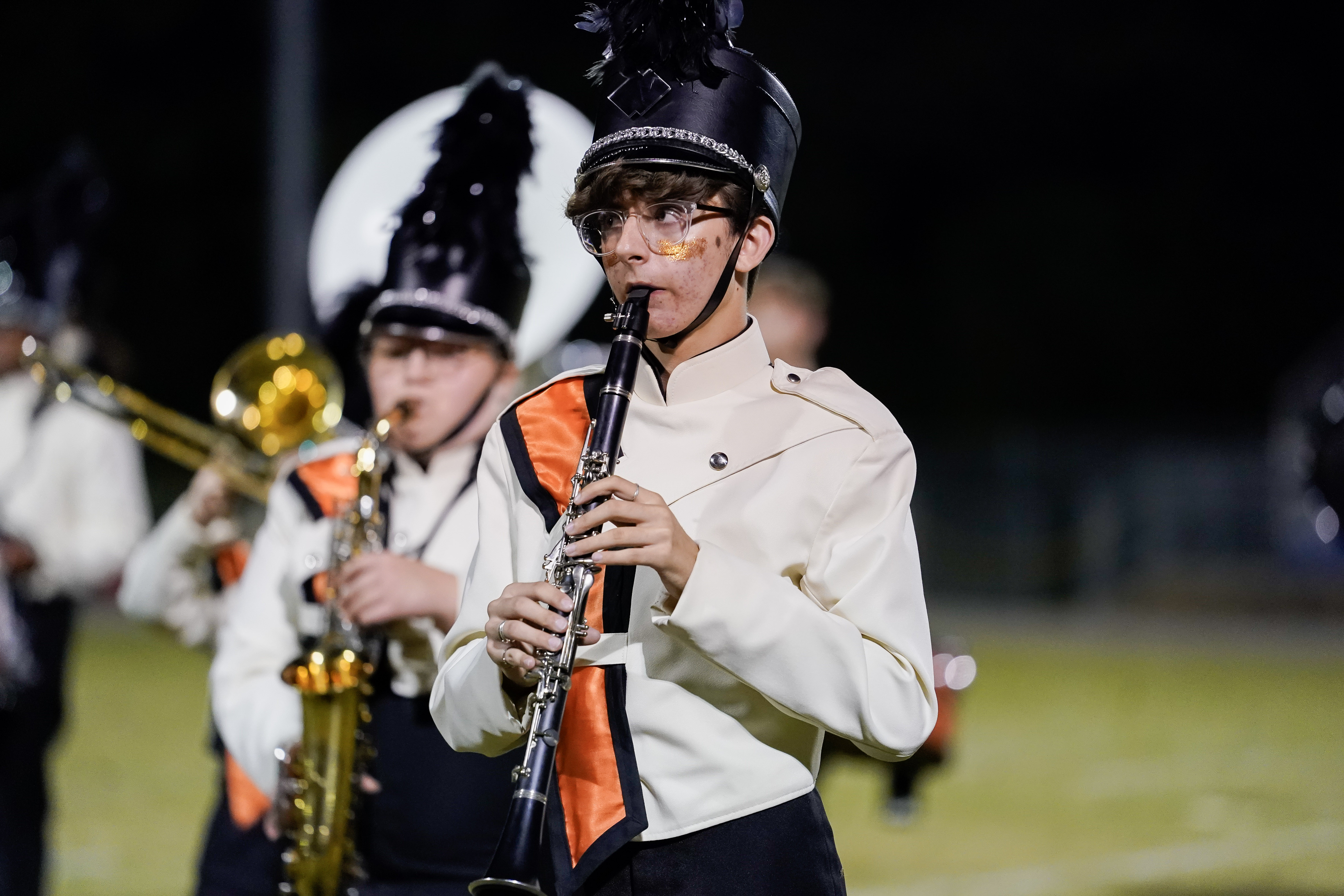 A marching band member in a white and black uniform with an orange sash plays a clarinet on a field at night. Other band members with instruments are visible in the background.