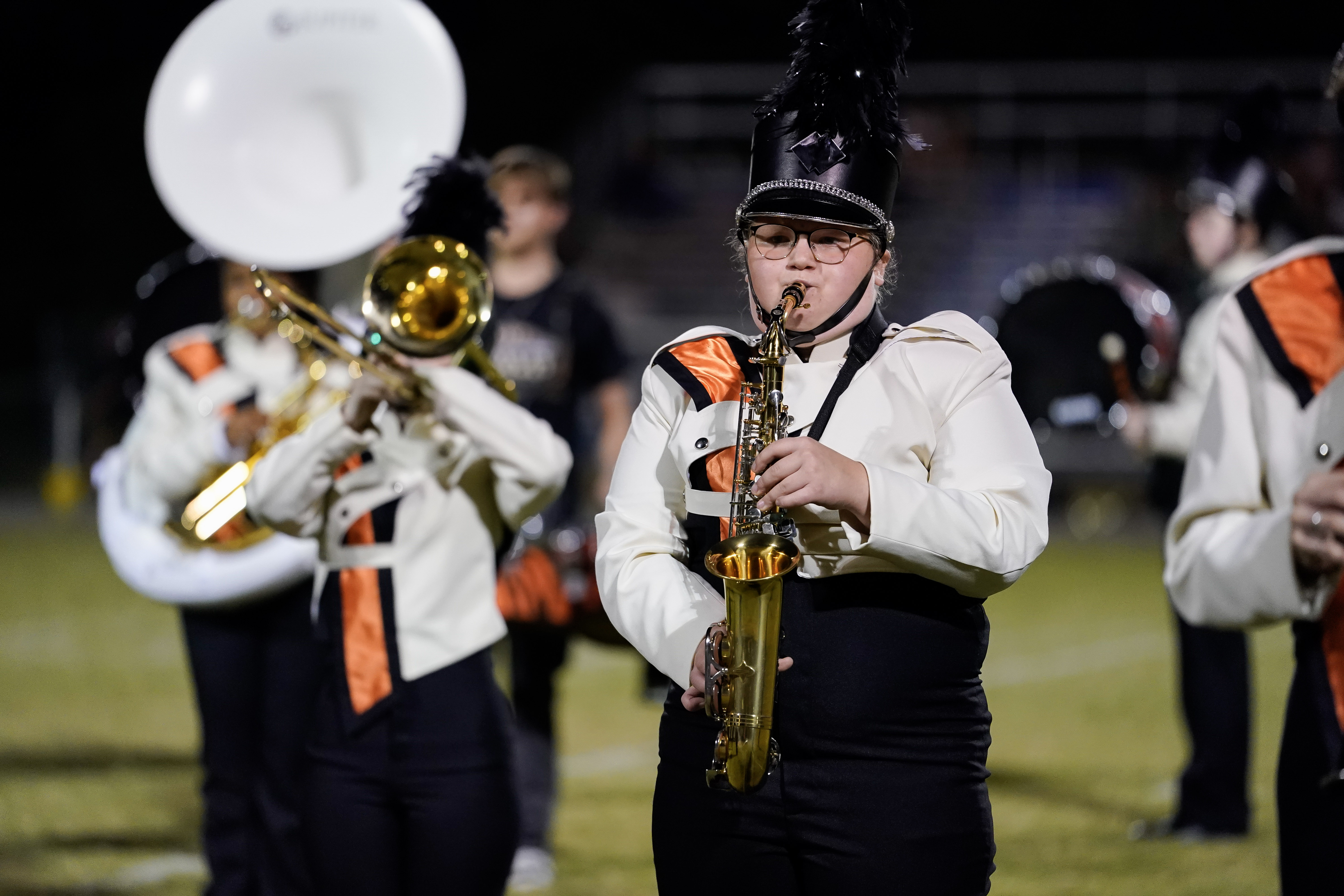 A marching band member in a white and black uniform with an orange accent plays a saxophone on a field at night. Other band members with brass instruments are visible in the background.