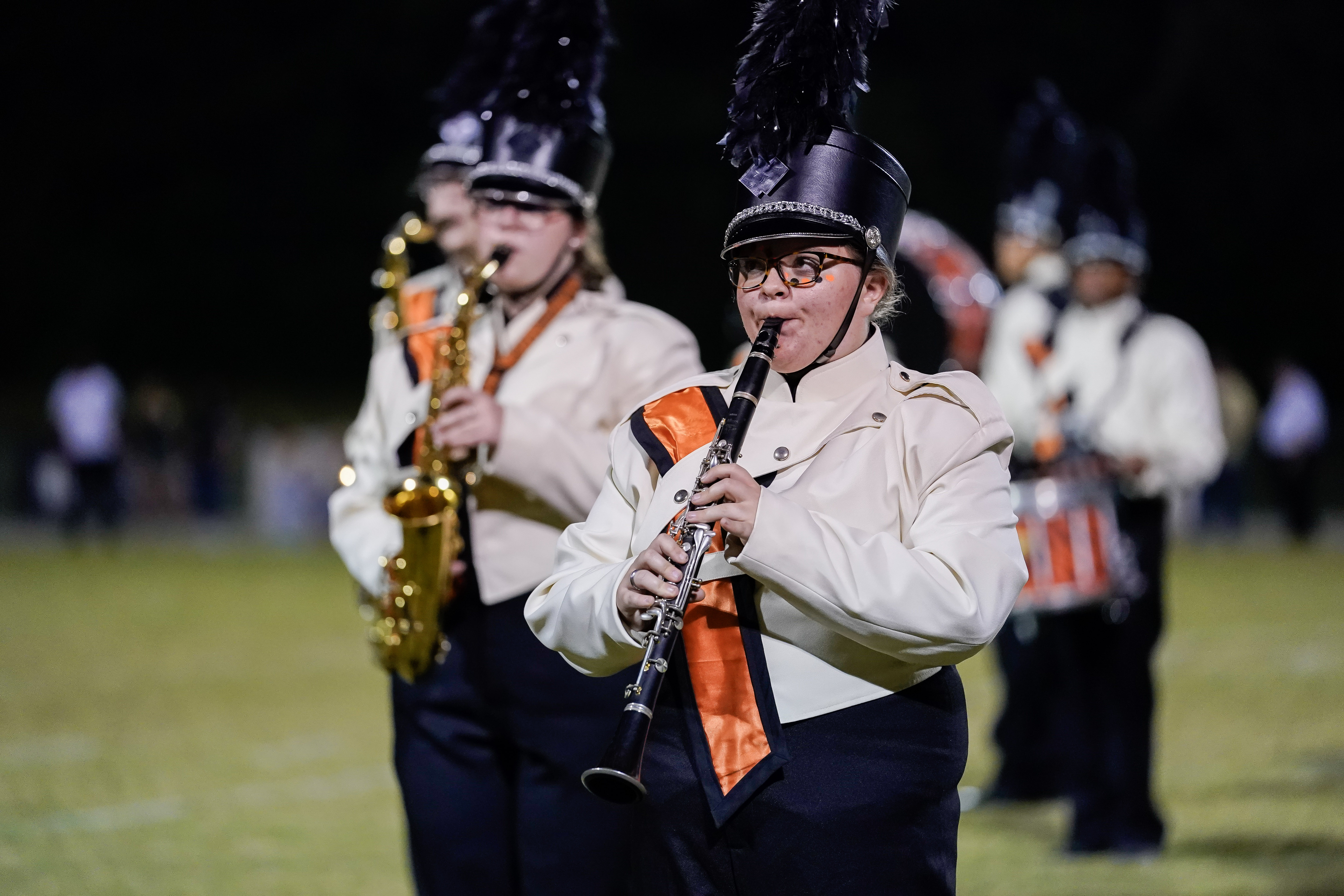 A marching band performs on a field at night. The focus is on a clarinet player in a uniform with a feathered hat and orange sash, with other band members playing instruments in the background.