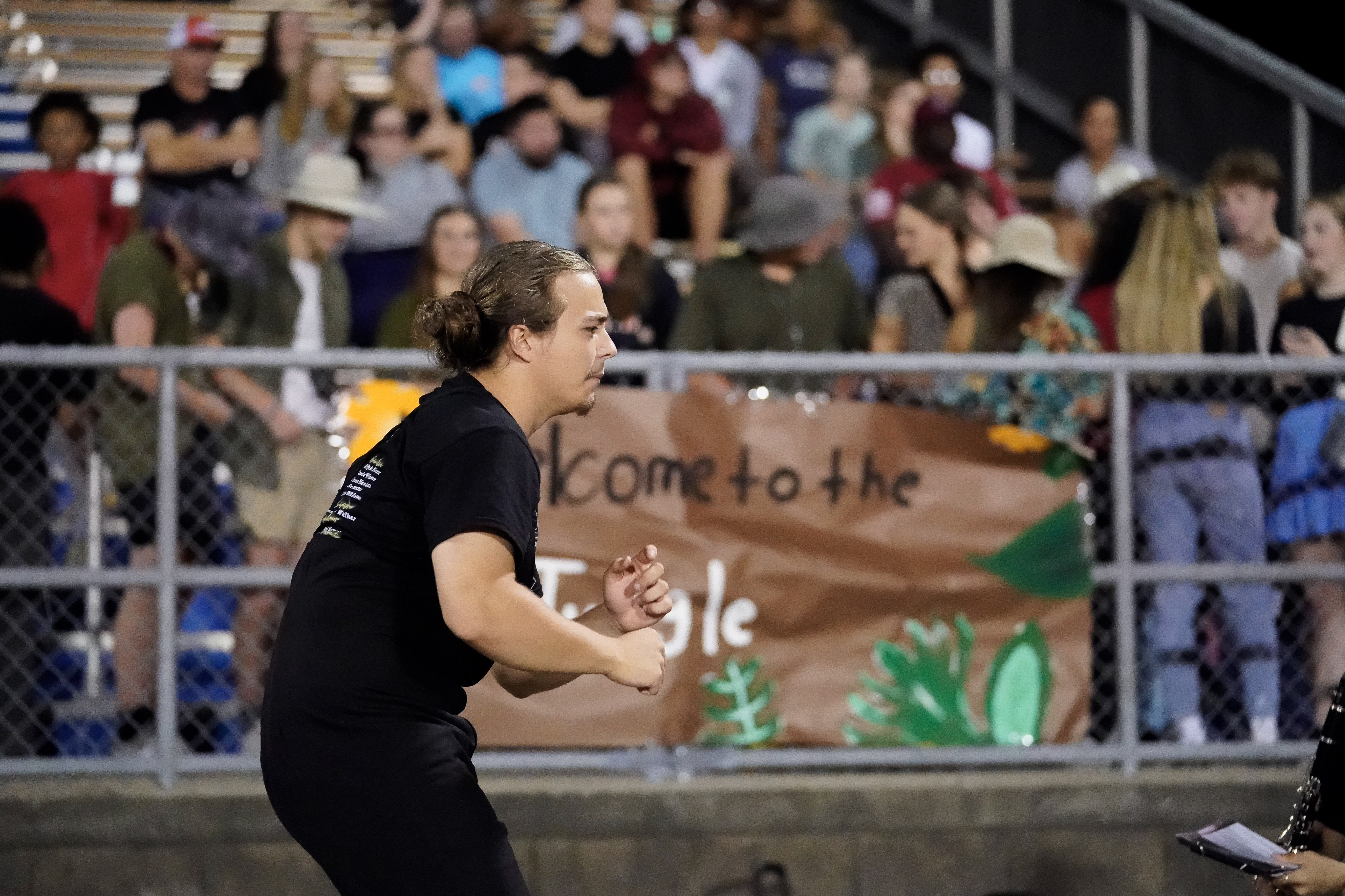 A man in a black shirt gestures energetically in front of bleachers filled with people. Behind him, a handmade sign reads Welcome to the Jungle. The crowd is watching attentively.