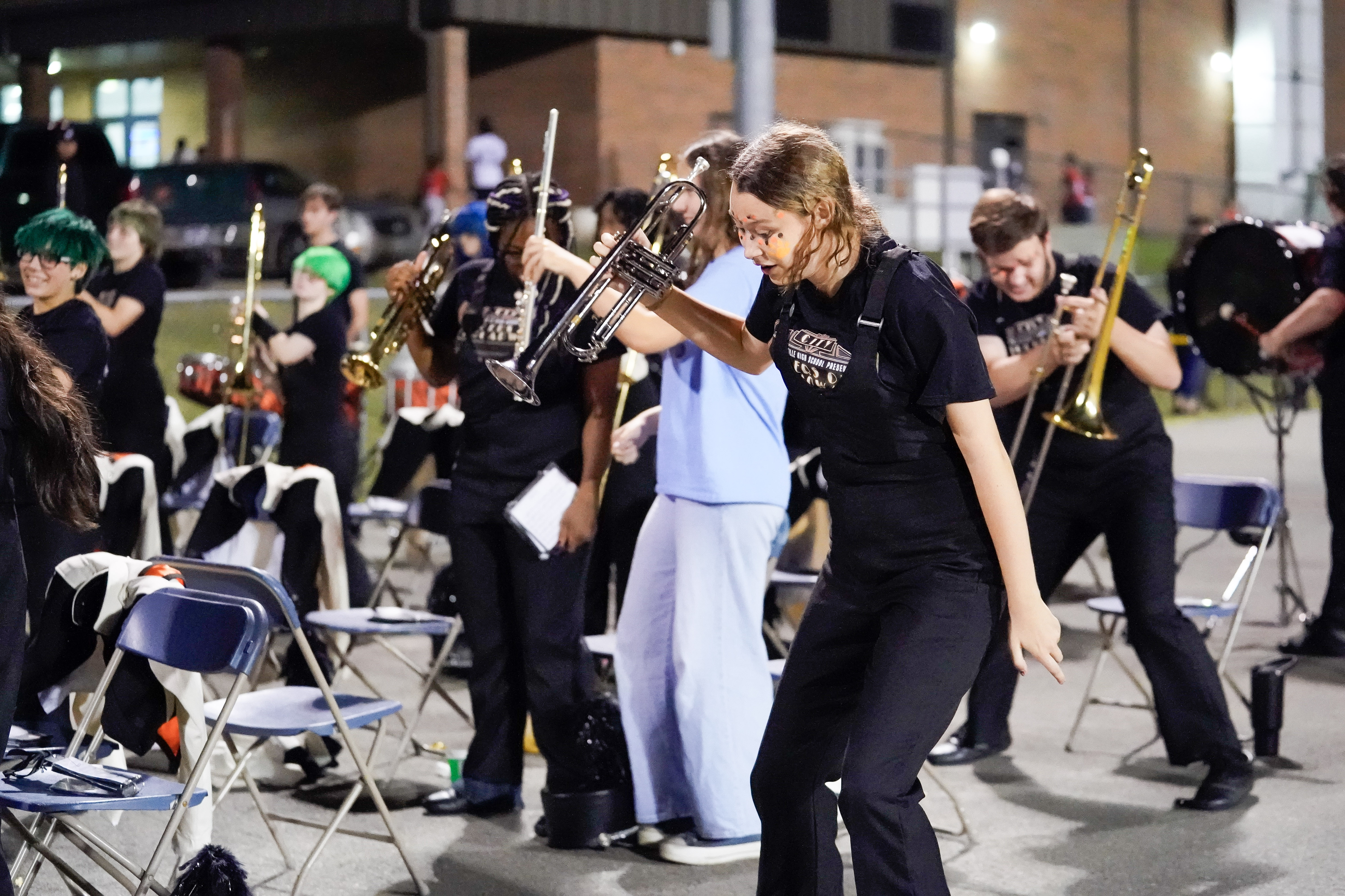 A group of marching band members, wearing black uniforms, play instruments and move energetically outdoors at night, with chairs and people in the background.