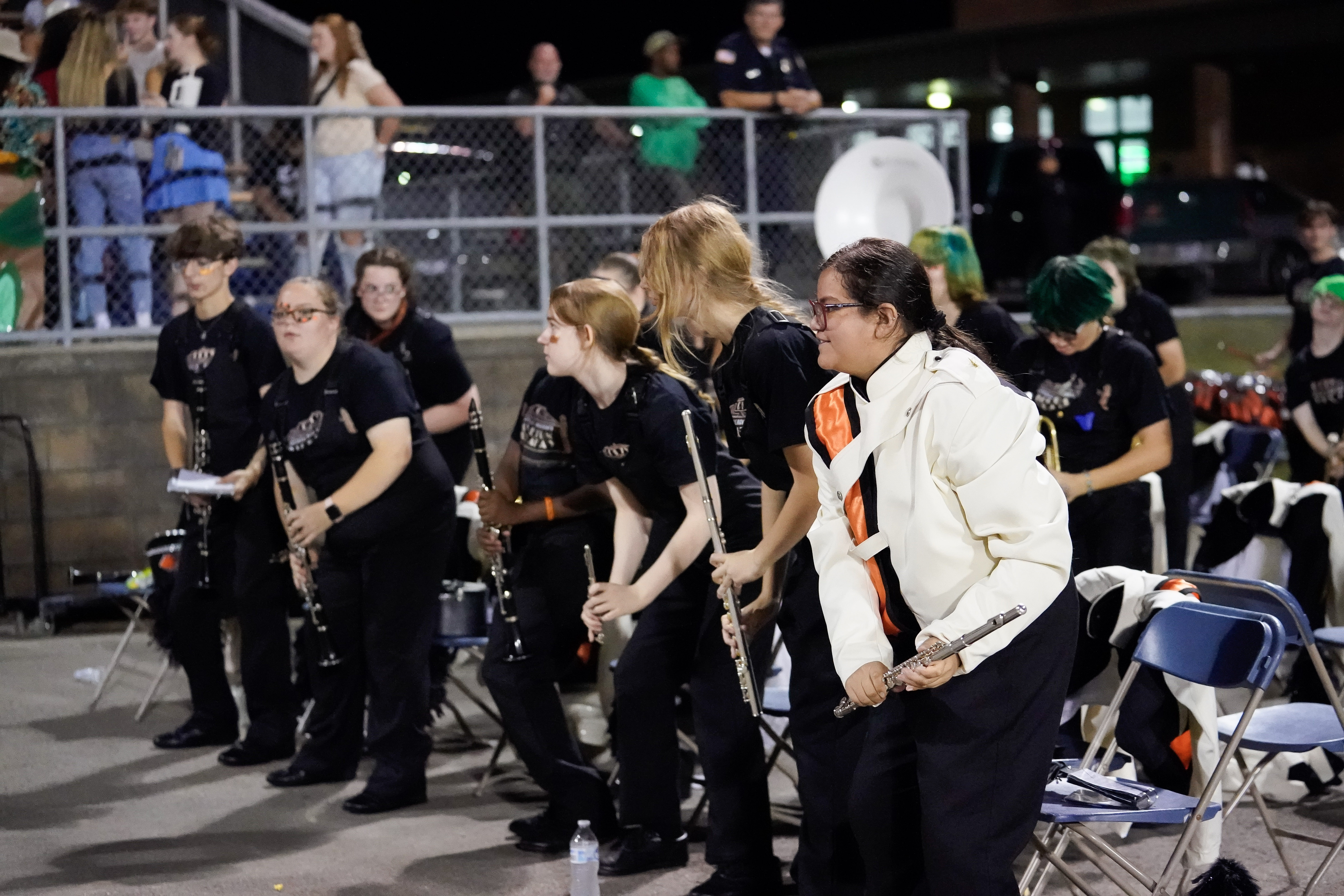 A group of band students in black uniforms, some holding clarinets, stand and cheer near folding chairs at night by a chain-link fence; one student wears a white marching band jacket.
