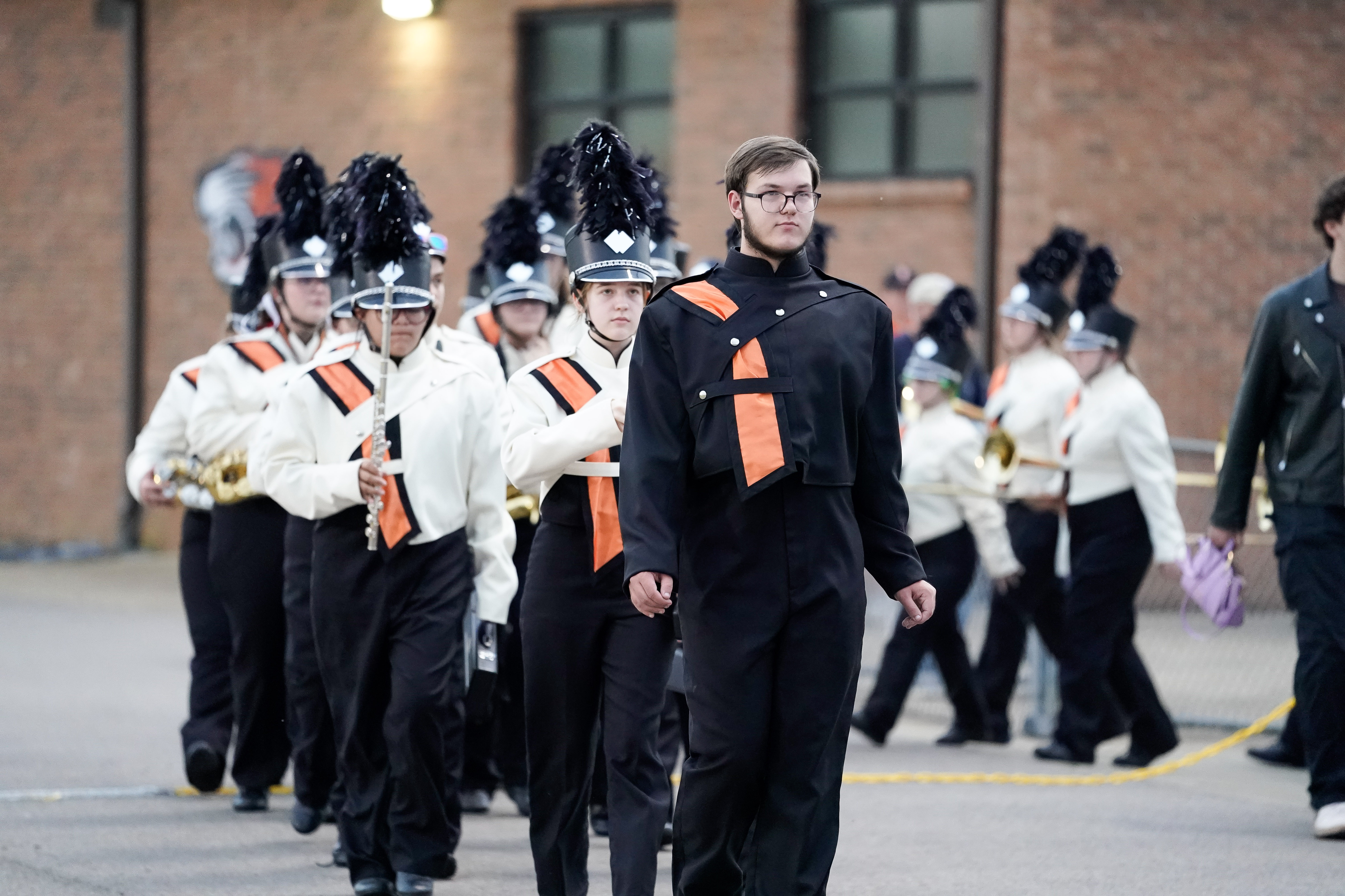 A marching band in black and white uniforms with feathered hats walks in formation outside, led by a drum major in predominantly black attire. The background shows a brick building and windows.