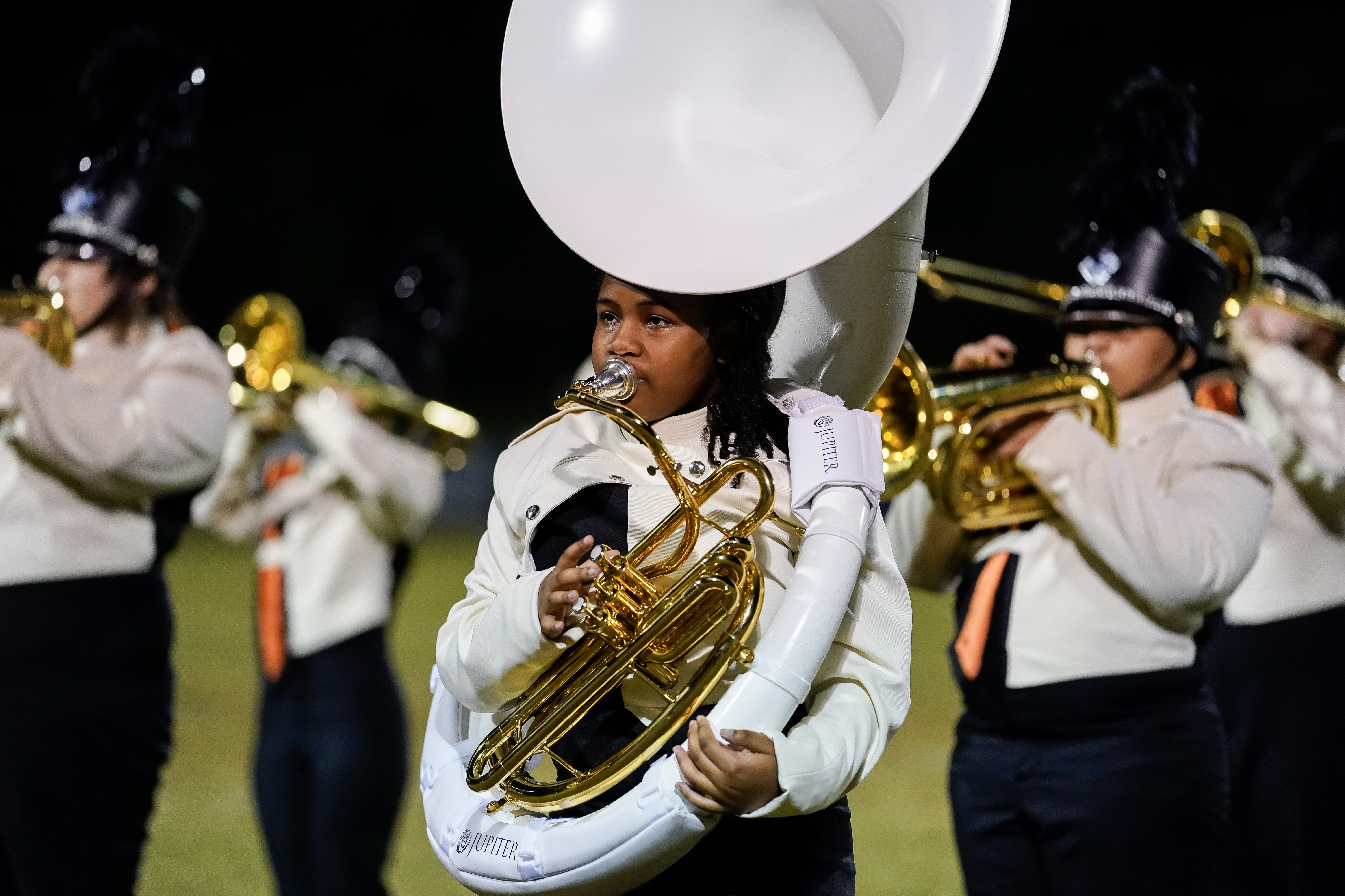 A young marching band member in uniform plays a white sousaphone, surrounded by other band members playing brass instruments during an outdoor performance at night.