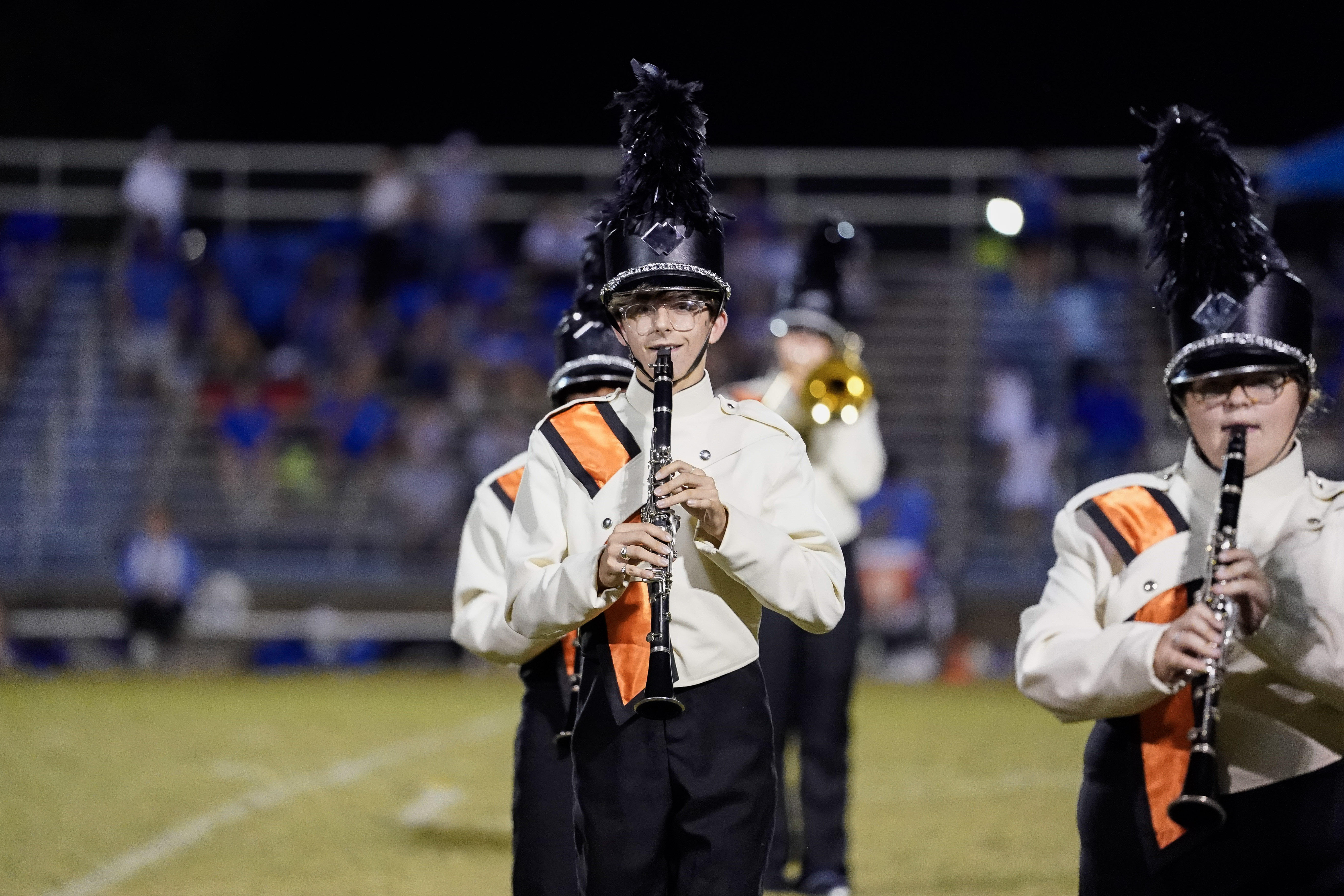 A marching band member in a white and black uniform with an orange sash plays a clarinet on a football field at night, with other band members and blurred stadium seats in the background.