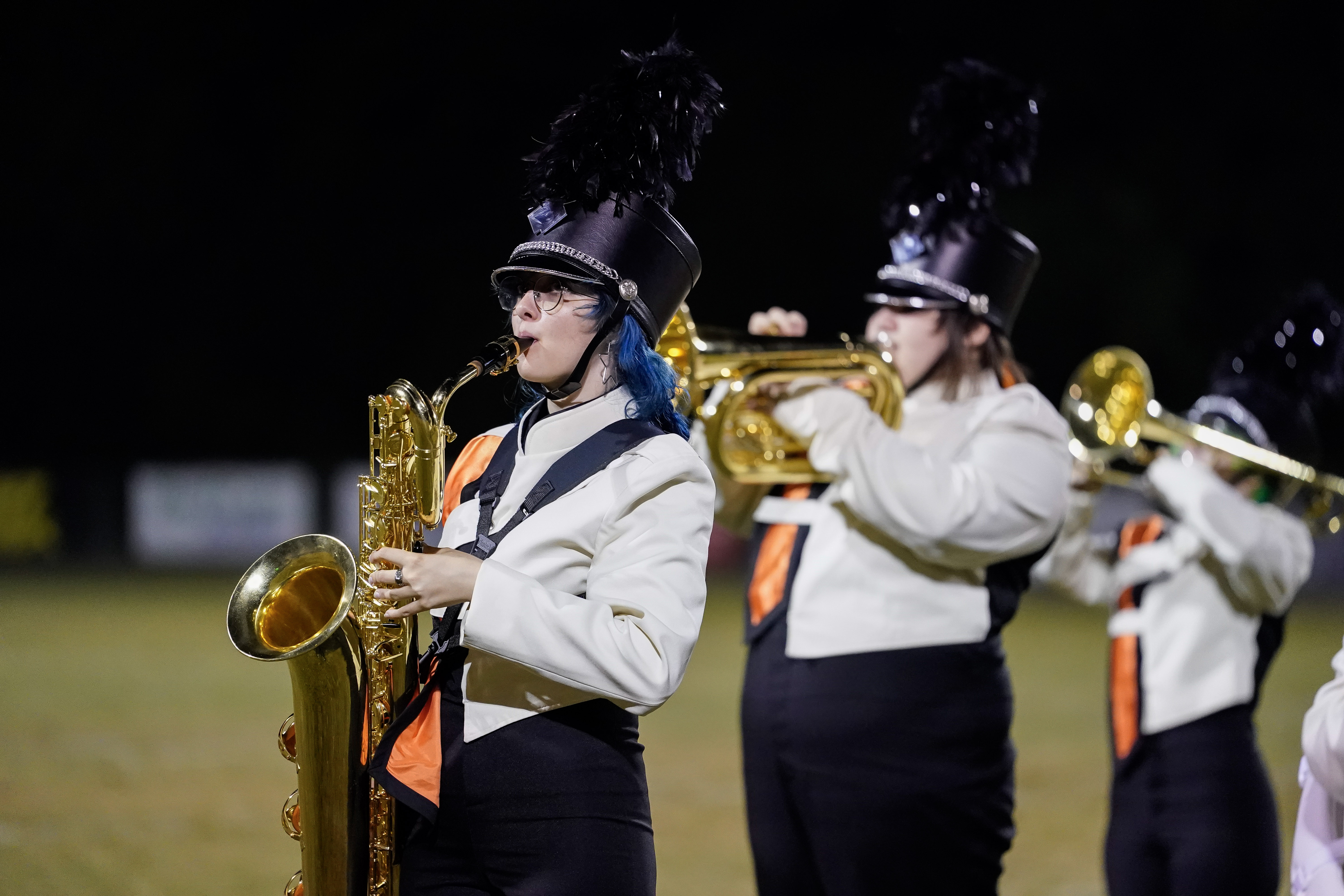 A marching band member in a white and black uniform with a feathered hat plays a saxophone at night, with other band members playing brass instruments in the background.