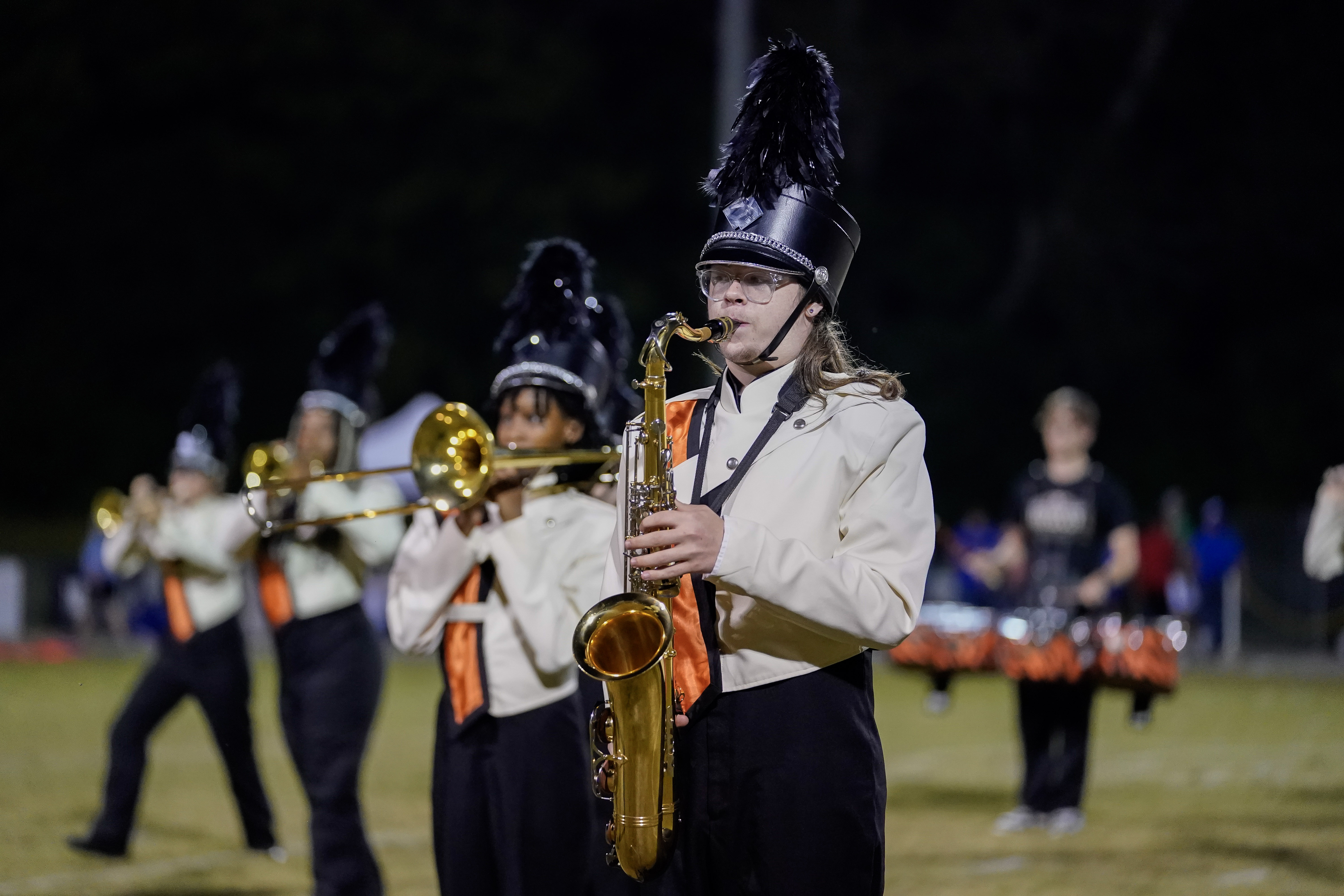 A marching band performs at night. In the foreground, a band member in a black hat with a feather plume plays a saxophone, while others behind play brass instruments and drums. They wear white jackets with orange and black details.