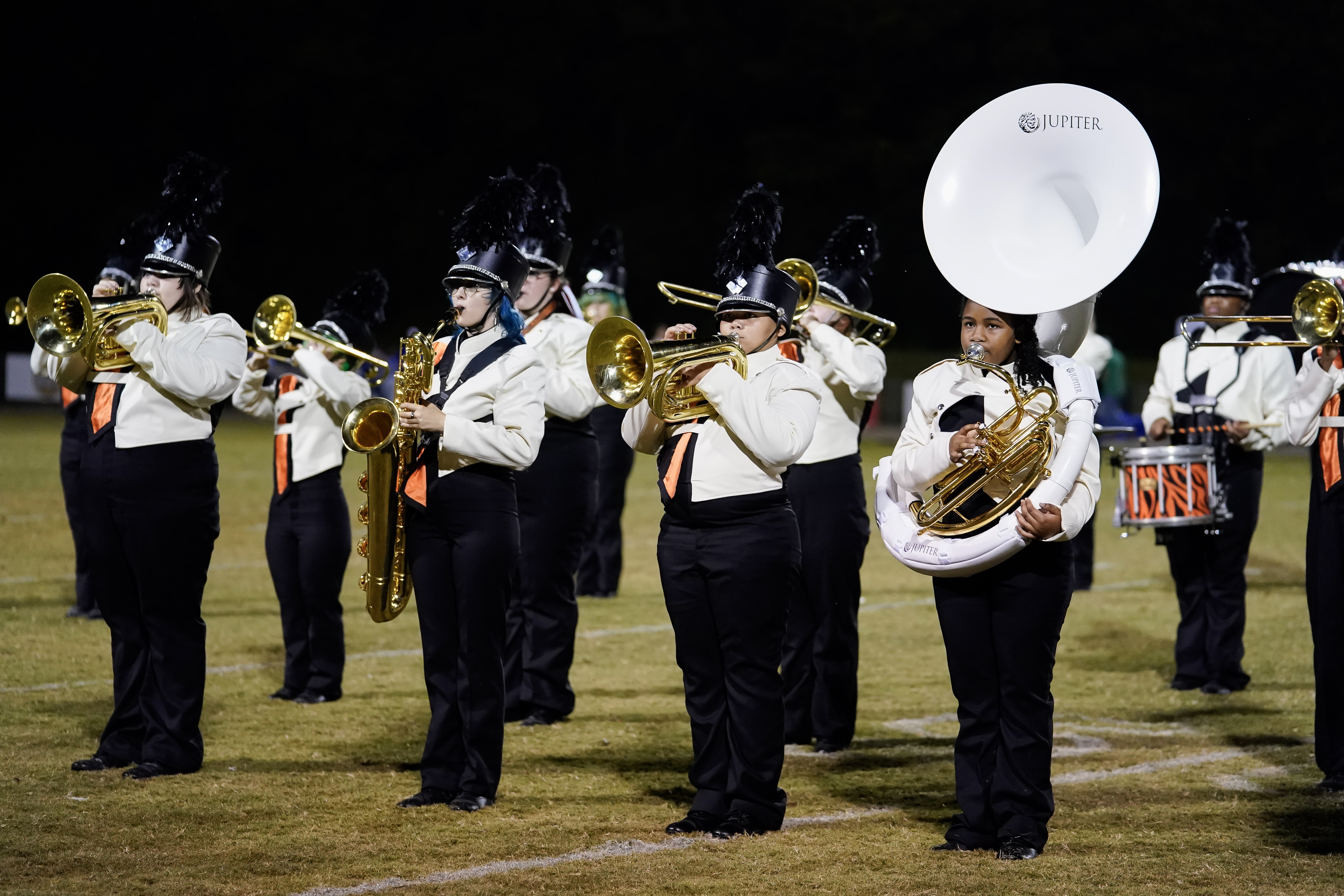 A marching band performs on a grassy field at night. Members wear white shirts, black pants, and tall black hats, playing brass and percussion instruments, including a sousaphone, trumpets, saxophones, and drums.