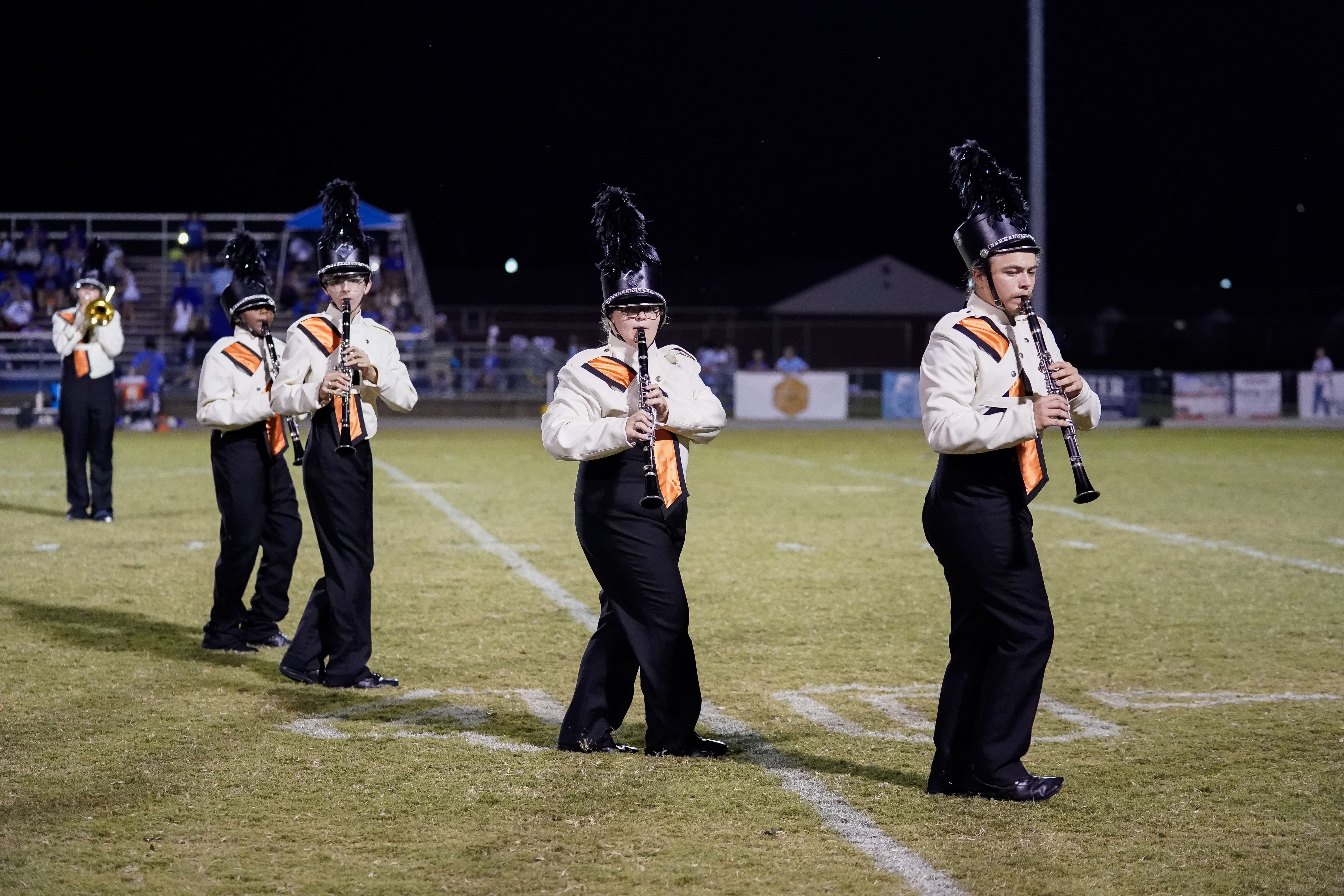 Four marching band members in uniforms and tall feathered hats play clarinets on a football field at night, performing in a diagonal line. Bleachers and a band are visible in the background.