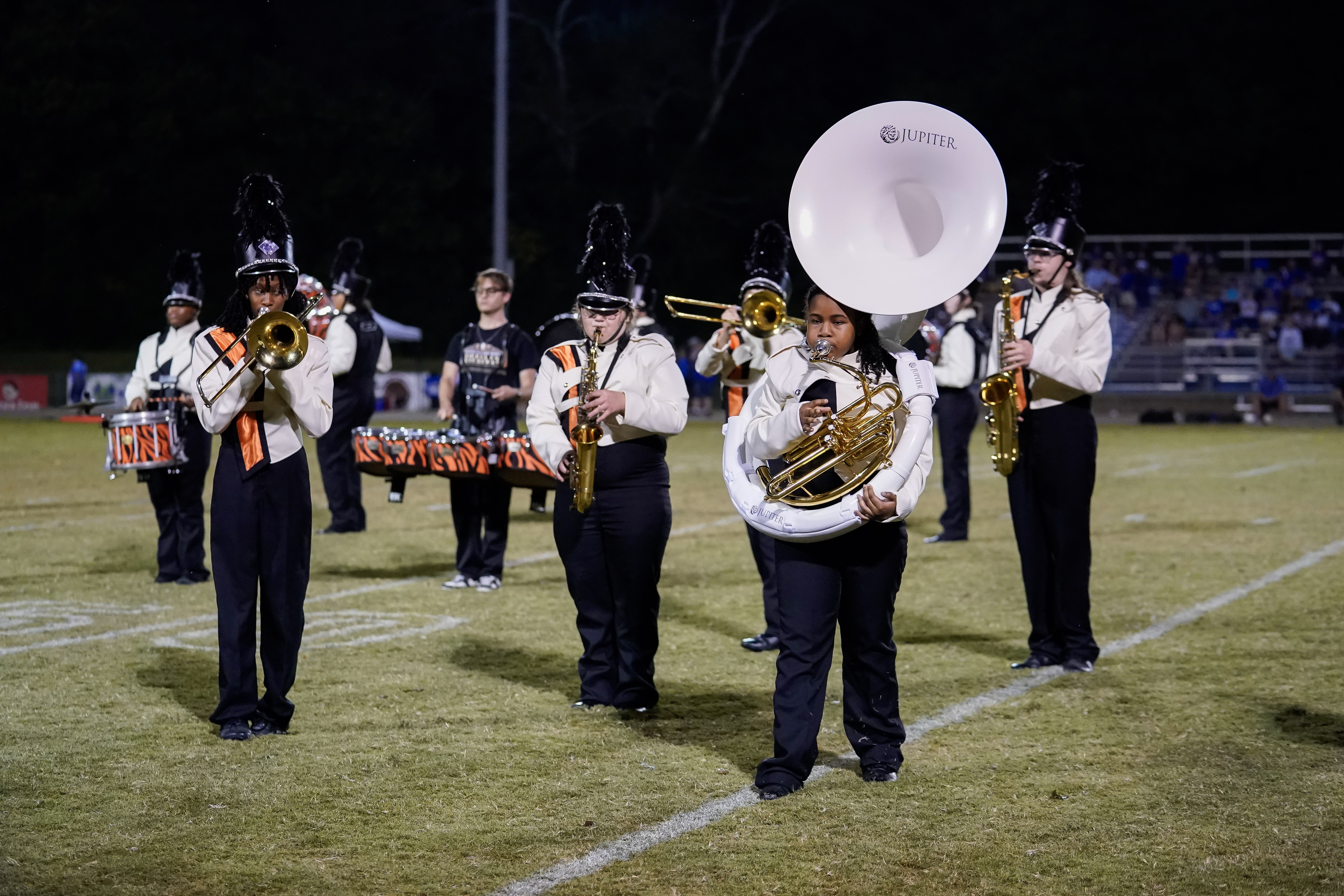 A high school marching band performs on a football field at night. Band members wear uniforms and hats with plumes, playing instruments like trombone, saxophone, sousaphone, and snare drums. Bleachers are visible in the background.