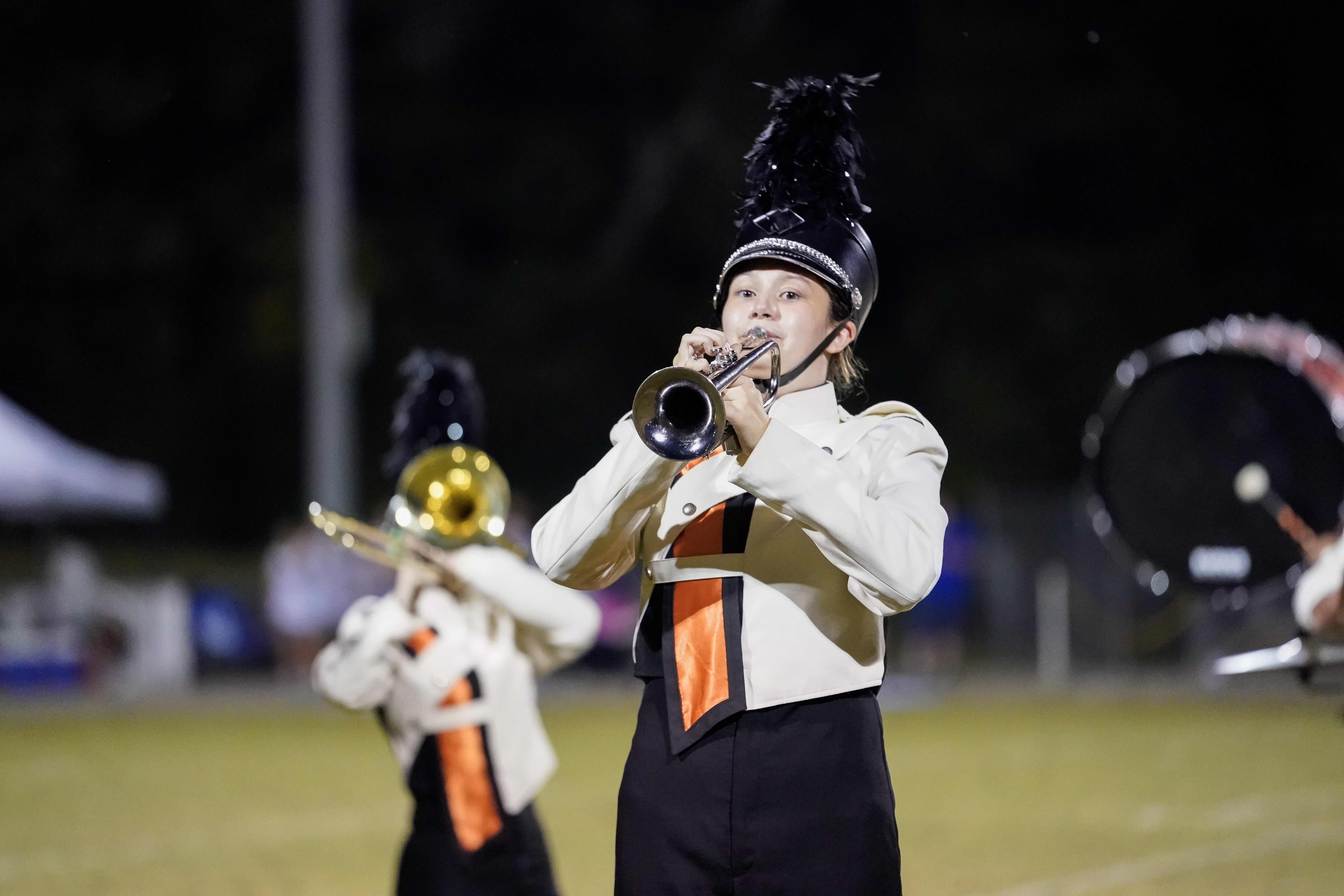 A marching band member in a white uniform and black hat plays the trumpet on a field at night, with another band member and a drummer visible in the background.