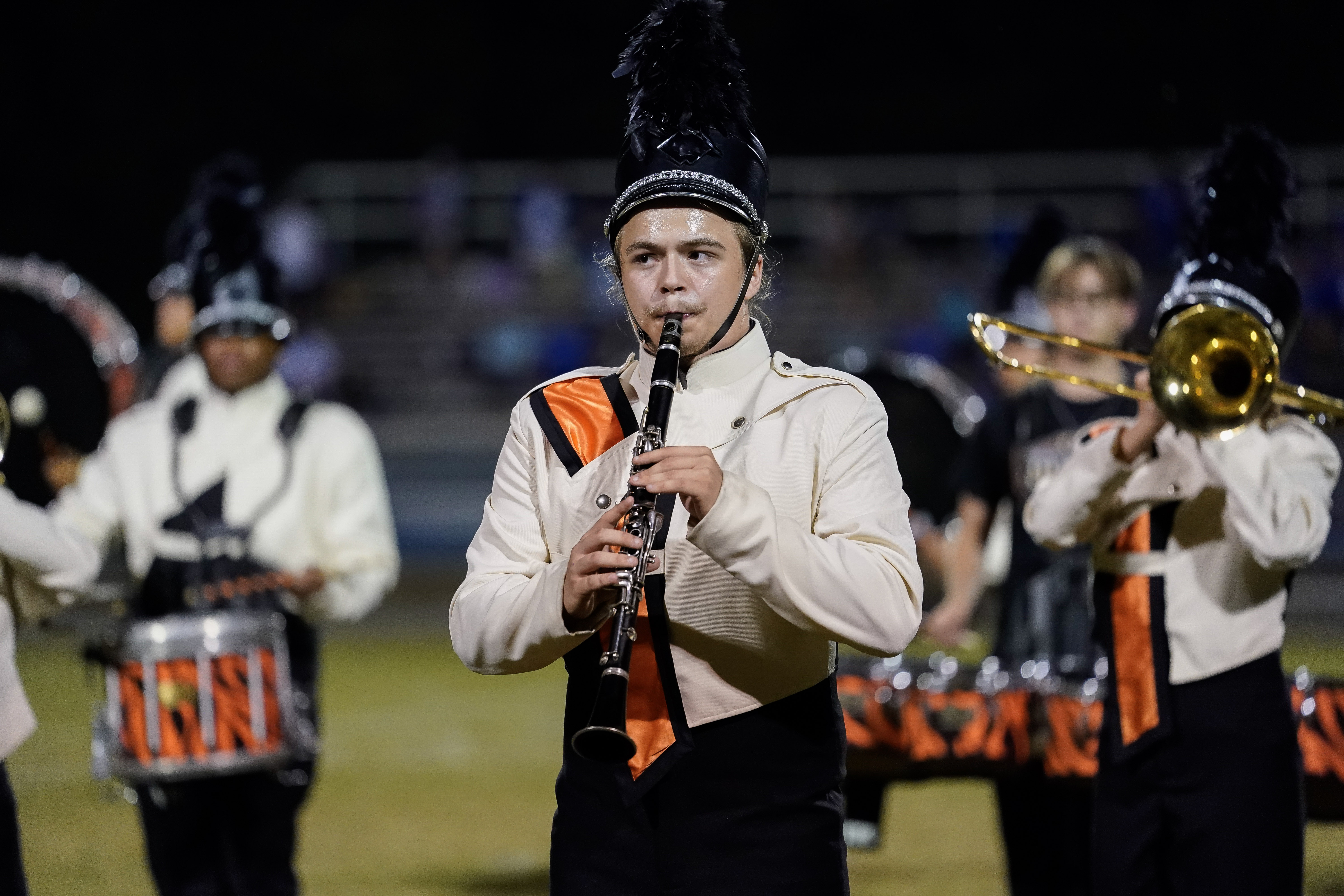 A marching band member in a cream and black uniform with an orange stripe plays a clarinet on a field at night. Other band members with instruments are visible in the background.