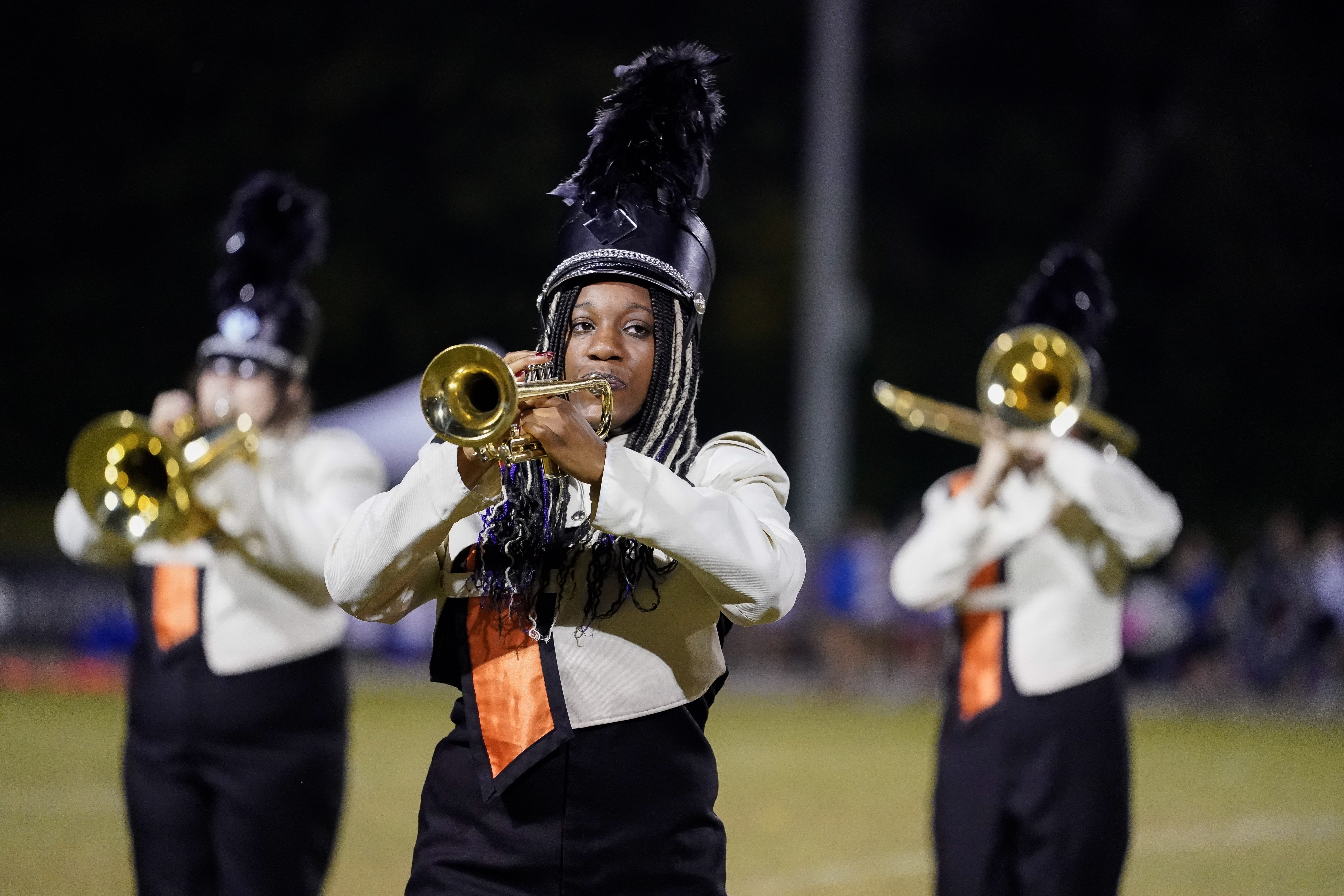 A marching band member in a black and white uniform with a feathered hat plays the trumpet outdoors, with two other band members playing trumpets in the background.