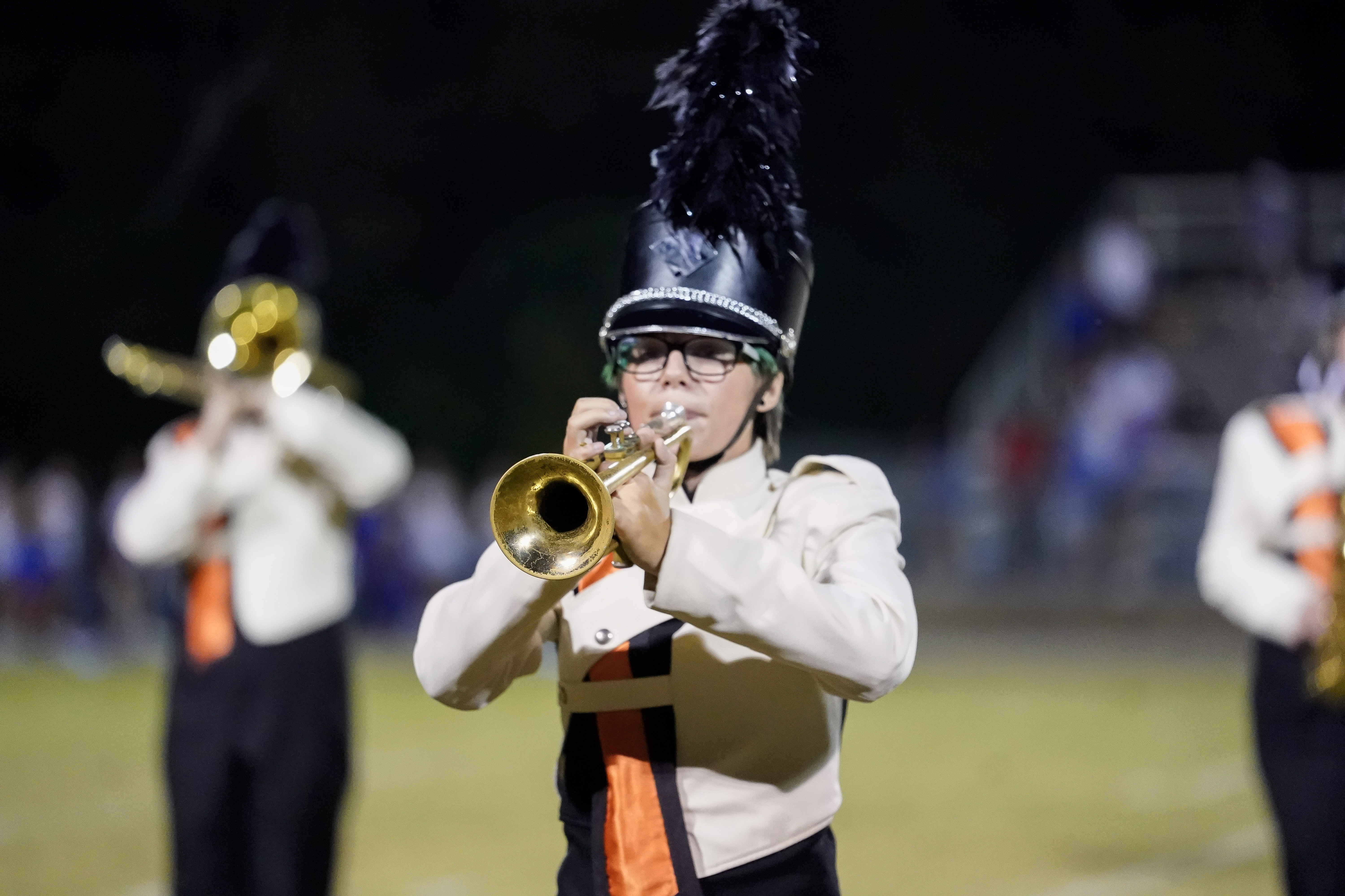 A marching band member in a cream and black uniform with a feathered hat plays a trumpet on a field at night. Other band members are visible in the blurred background.