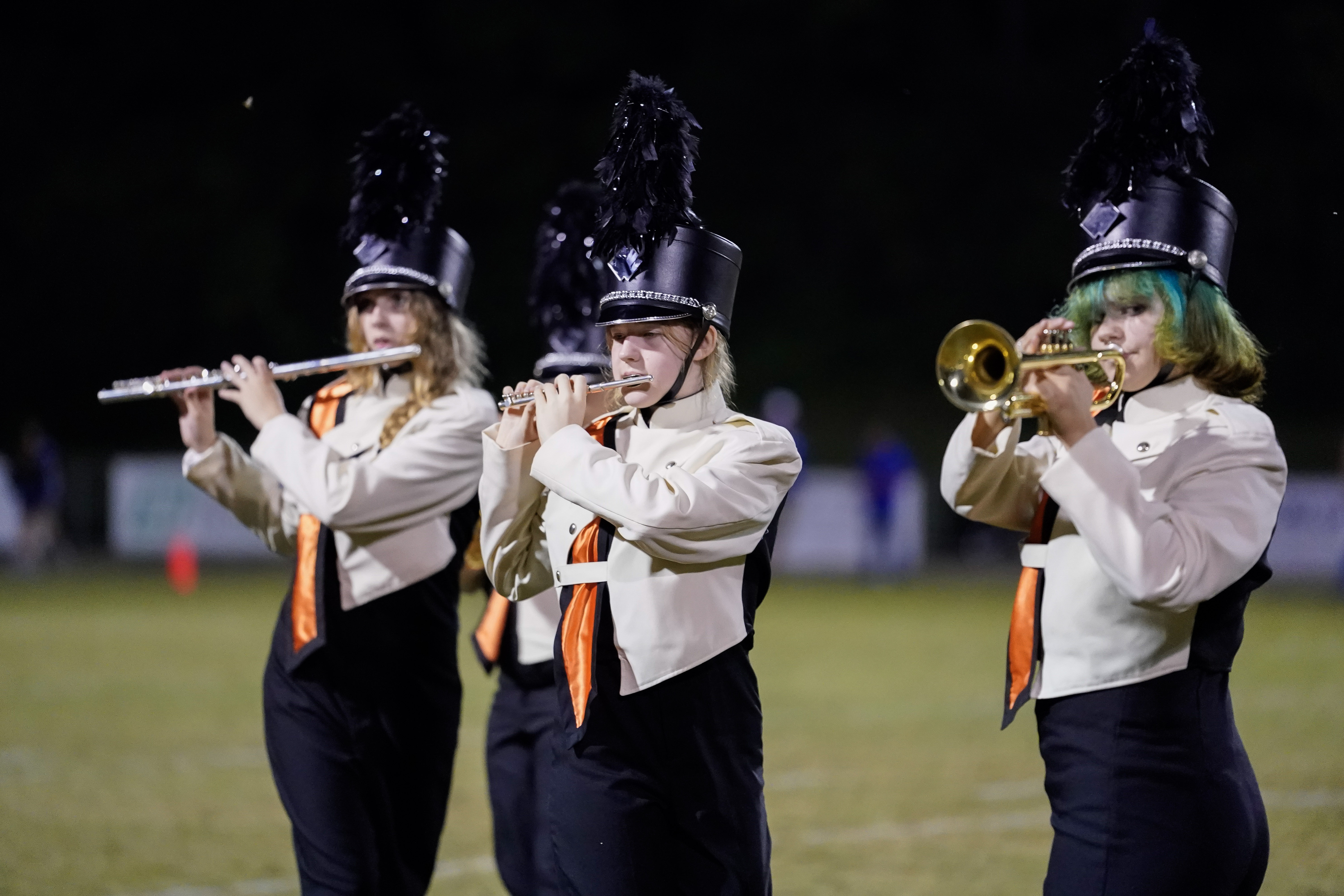 Three marching band members in uniforms and feathered hats play instruments—a flute, a clarinet, and a trumpet—on a field at night. The background is out of focus.