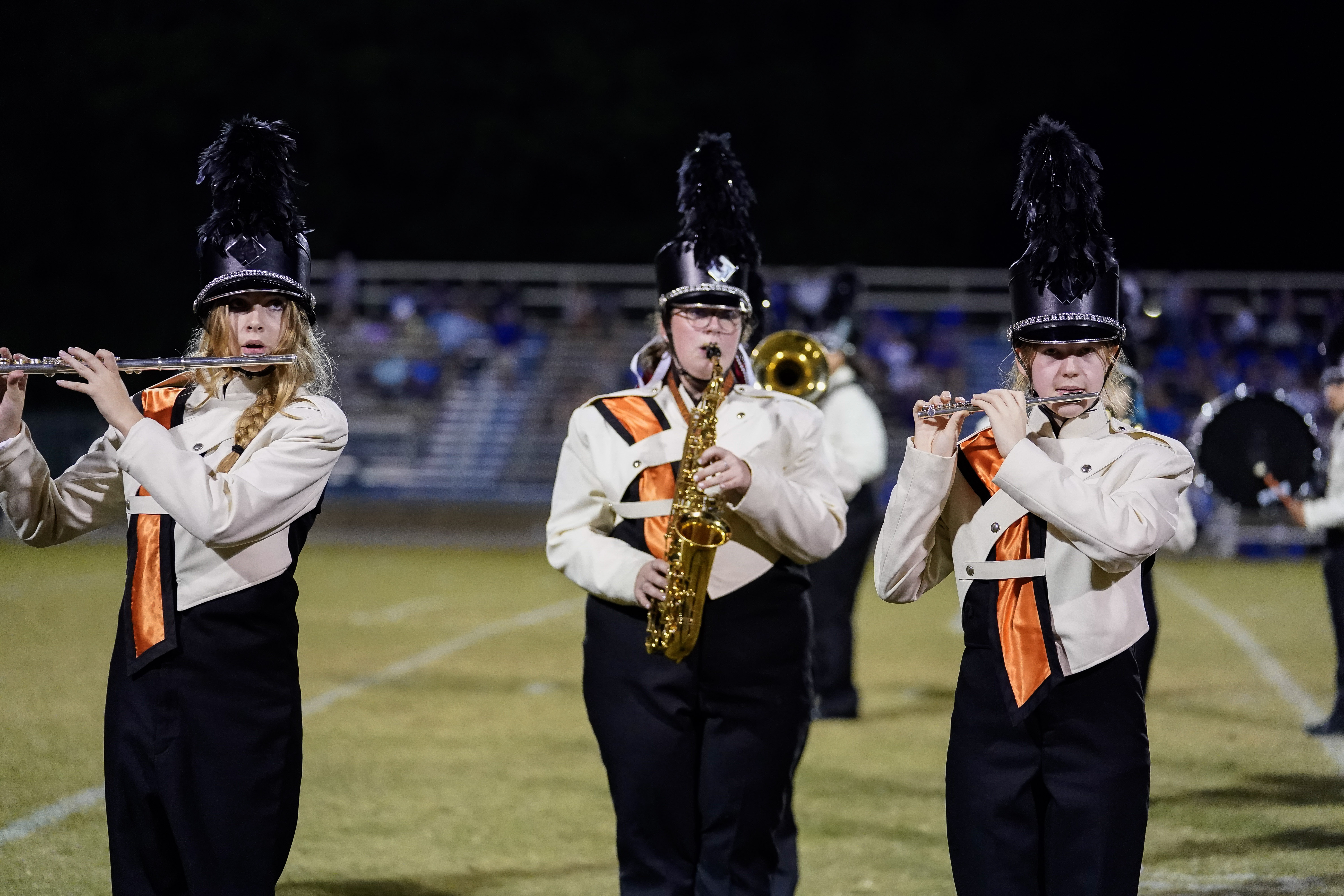 Three marching band members in uniform play instruments on a football field at night: a flute, a saxophone, and a clarinet. Bleachers and spectators are visible in the blurred background.