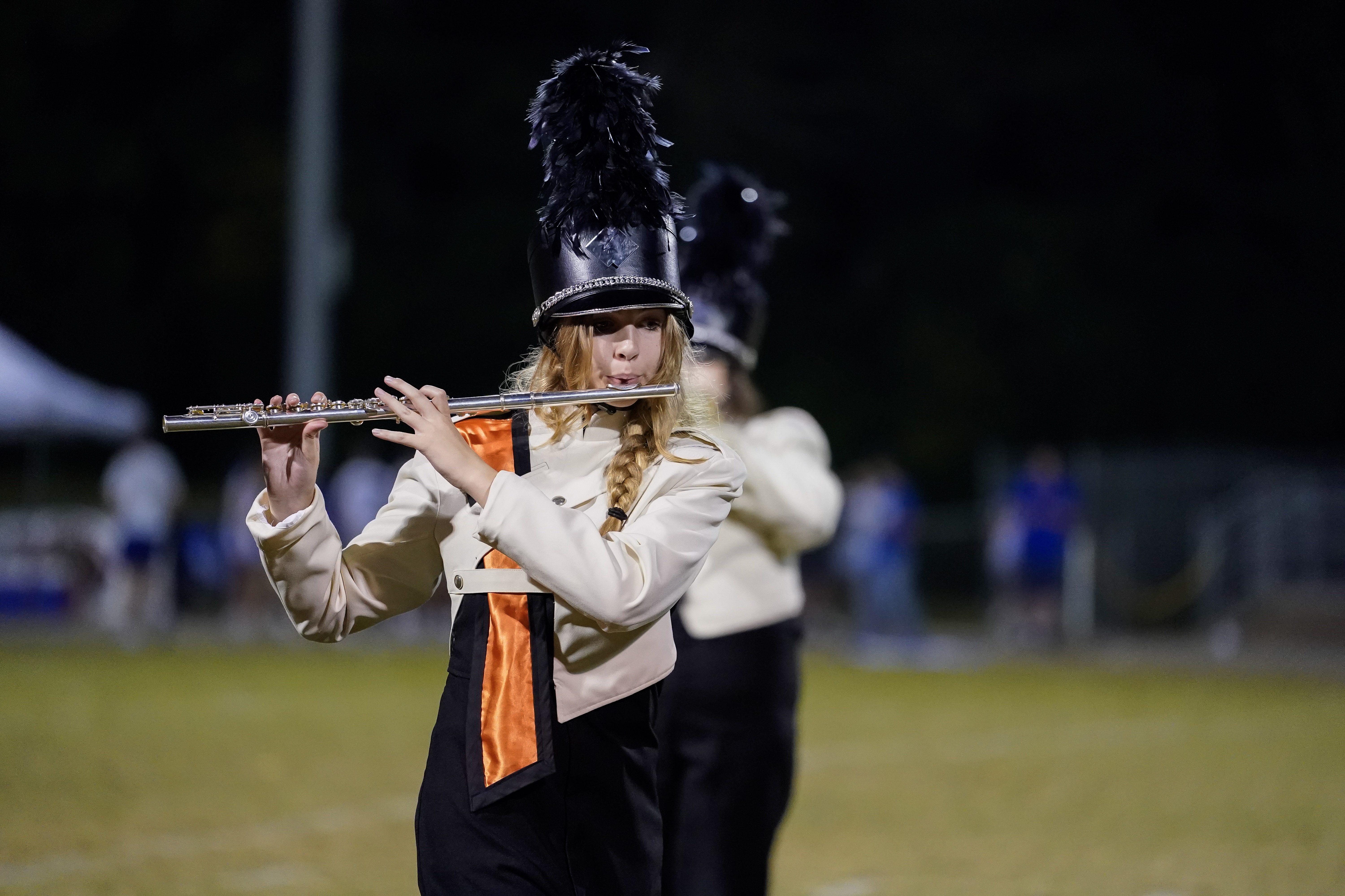 A marching band member in a black and white uniform with an orange sash plays the flute outdoors at night. She wears a tall hat with a black plume and stands on a field with blurred people in the background.