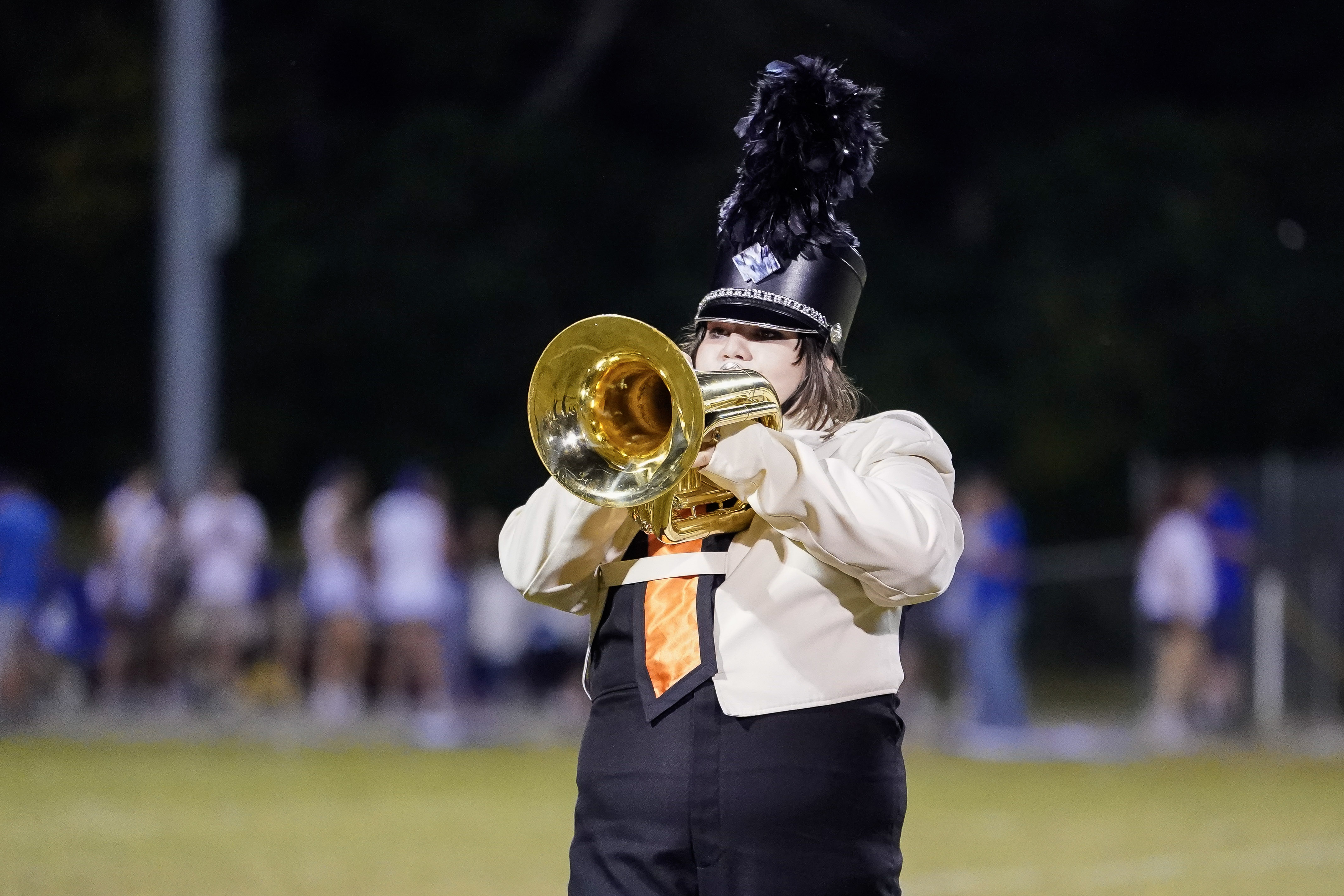 A marching band member in a black and cream uniform with a tall feathered hat plays a brass instrument, likely a mellophone, outdoors at night. A blurred crowd and field are visible in the background.