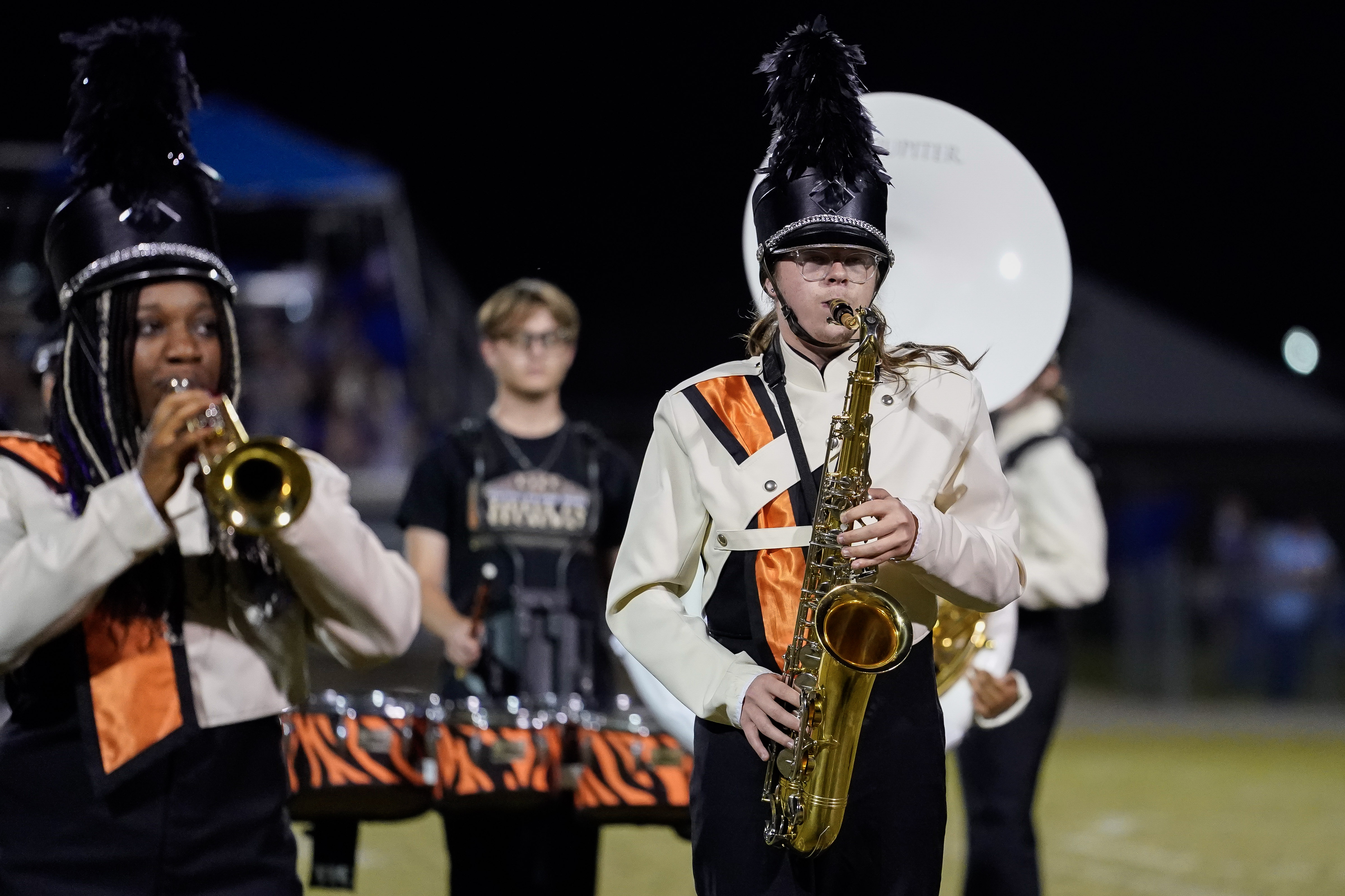 Two marching band members in black and white uniforms with orange accents perform at night; one plays a saxophone and the other plays a trombone on a football field.
