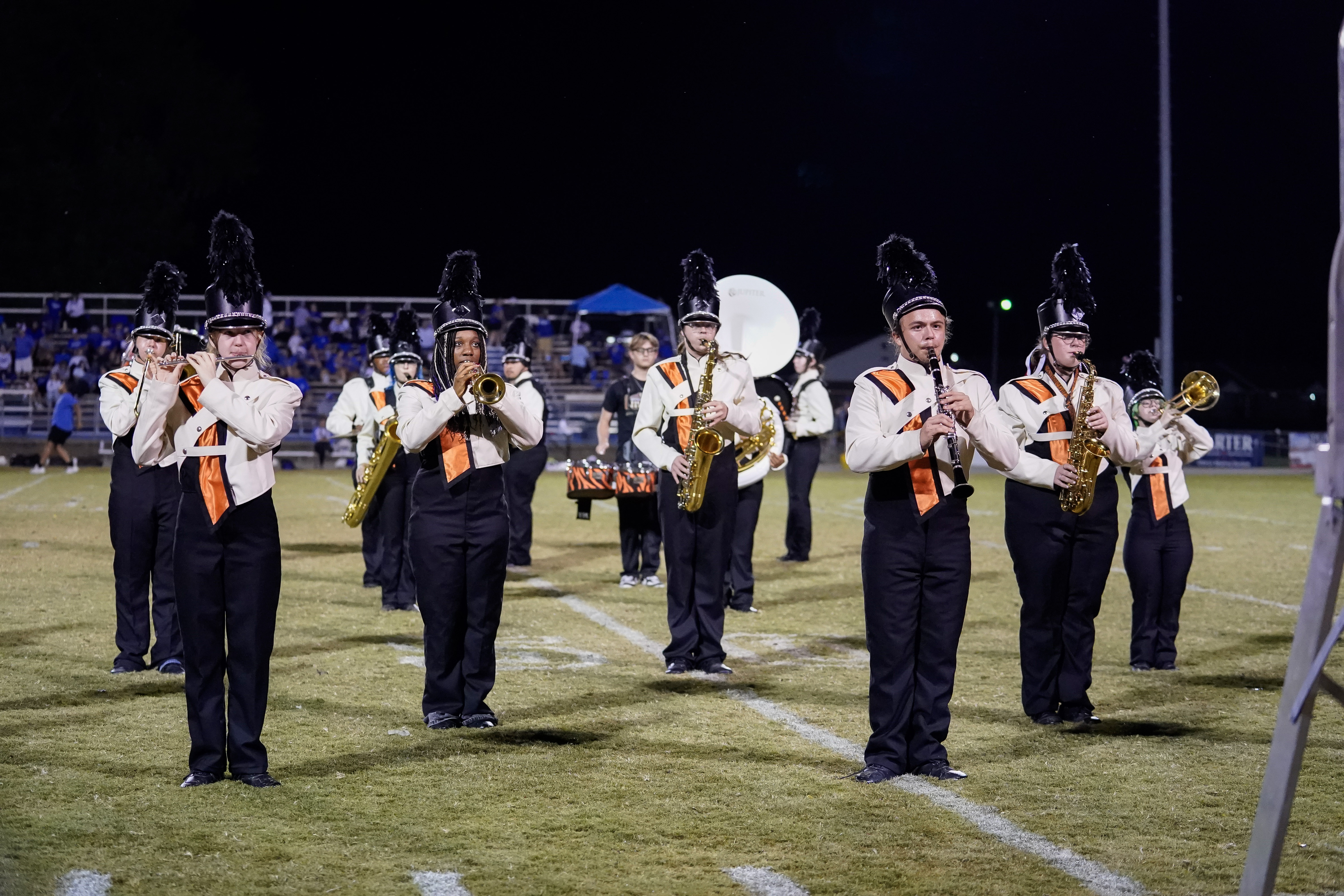 A high school marching band performs on a football field at night, with musicians wearing black and white uniforms and tall feathered hats, playing various brass and woodwind instruments. Spectators are visible in the background.