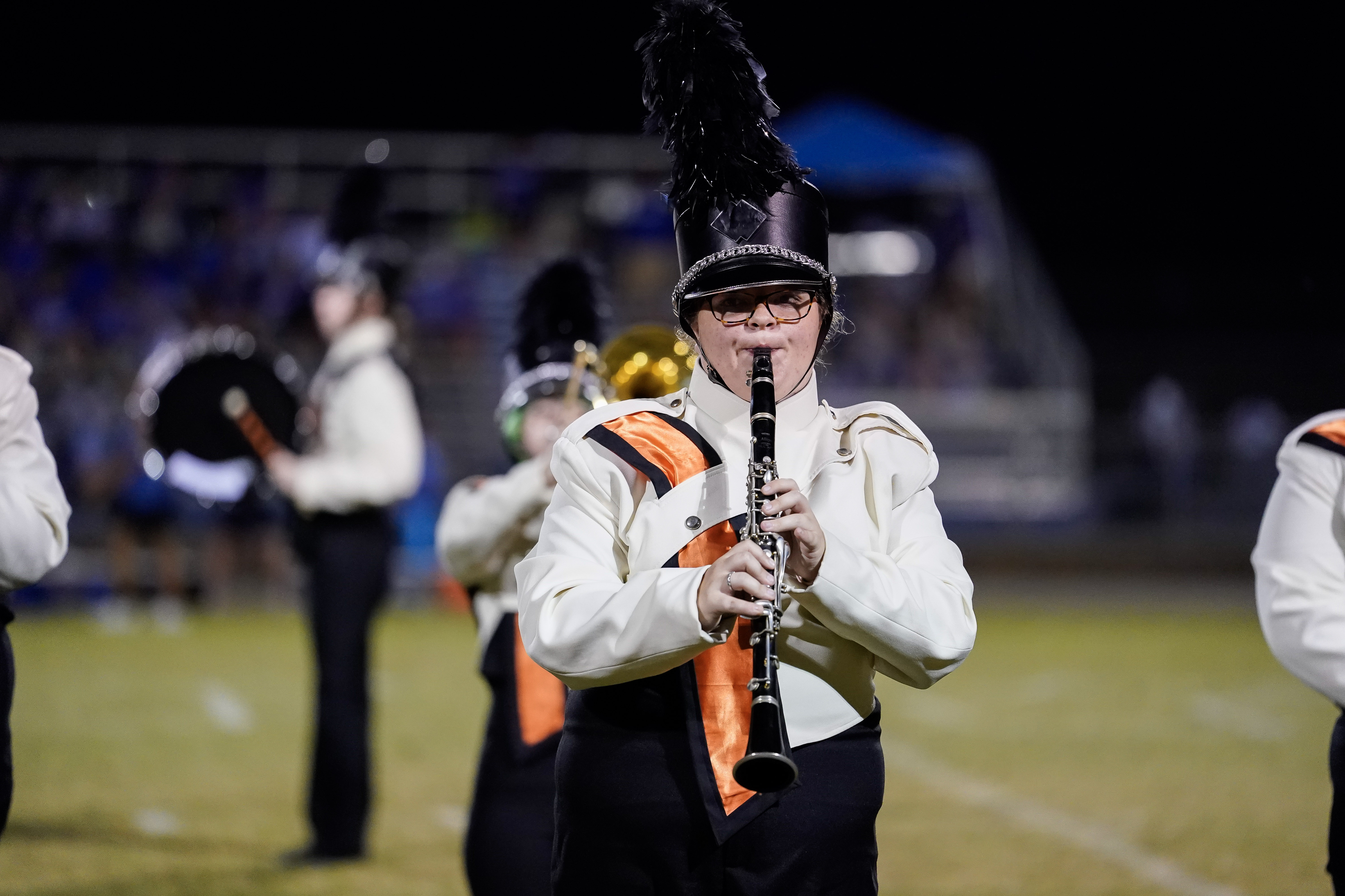 A marching band member in uniform and a feathered hat plays the clarinet on a field at night, with other band members performing in the background.