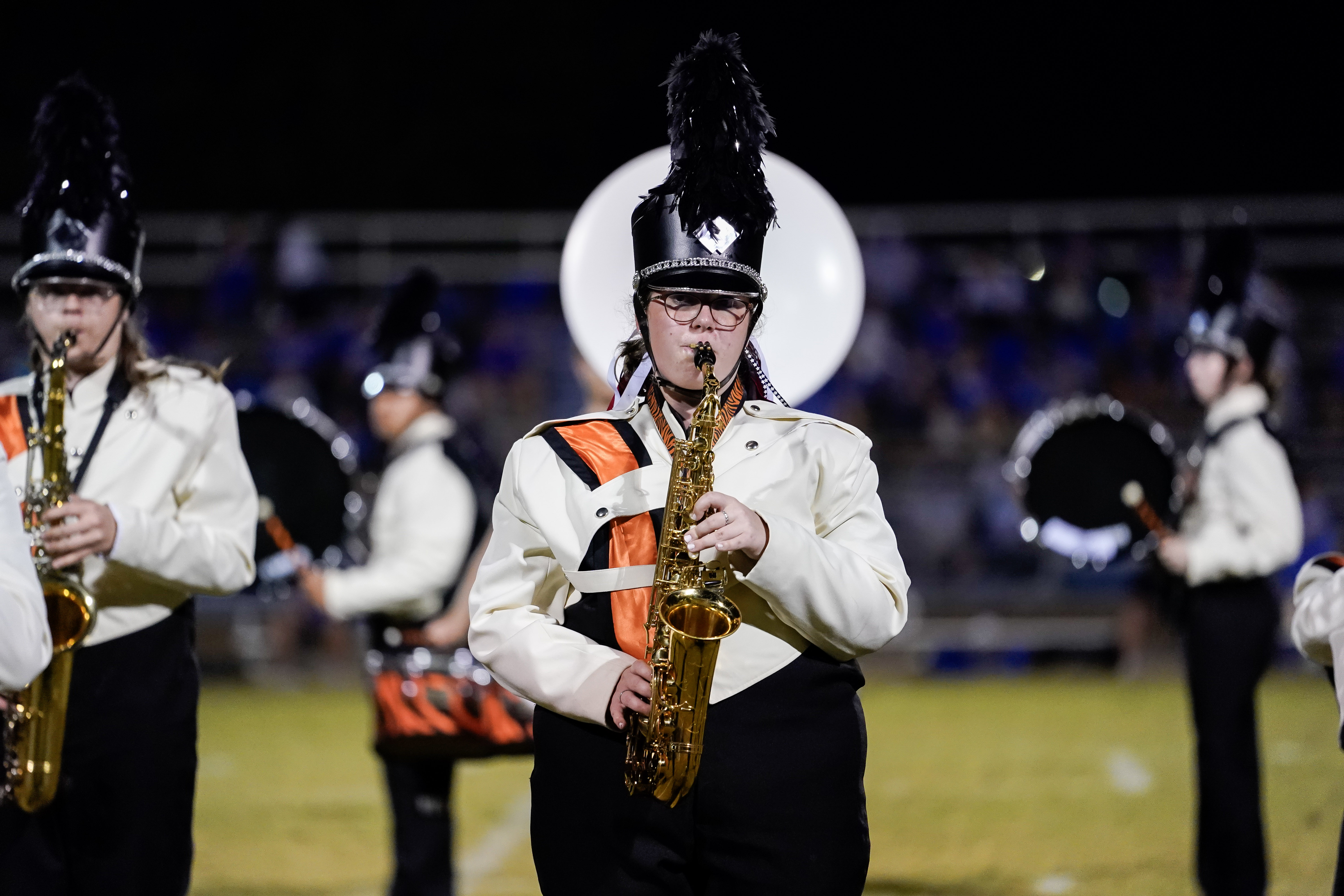 A marching band member in a white and black uniform with an orange stripe plays a saxophone on a field at night, surrounded by other band members in similar uniforms.