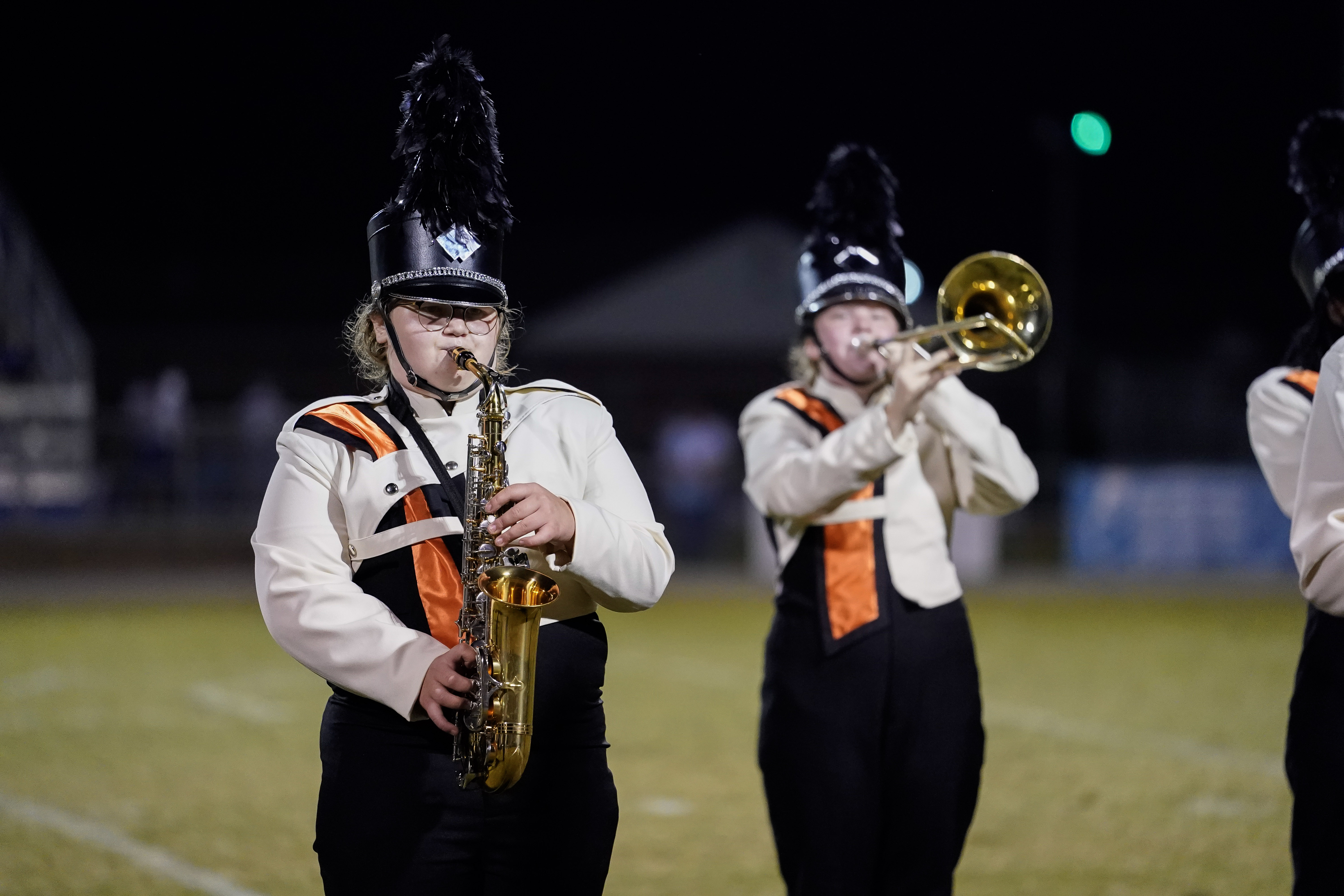 Two marching band members in black and white uniforms with orange accents perform at night; one plays a saxophone and the other plays a trombone on a football field.