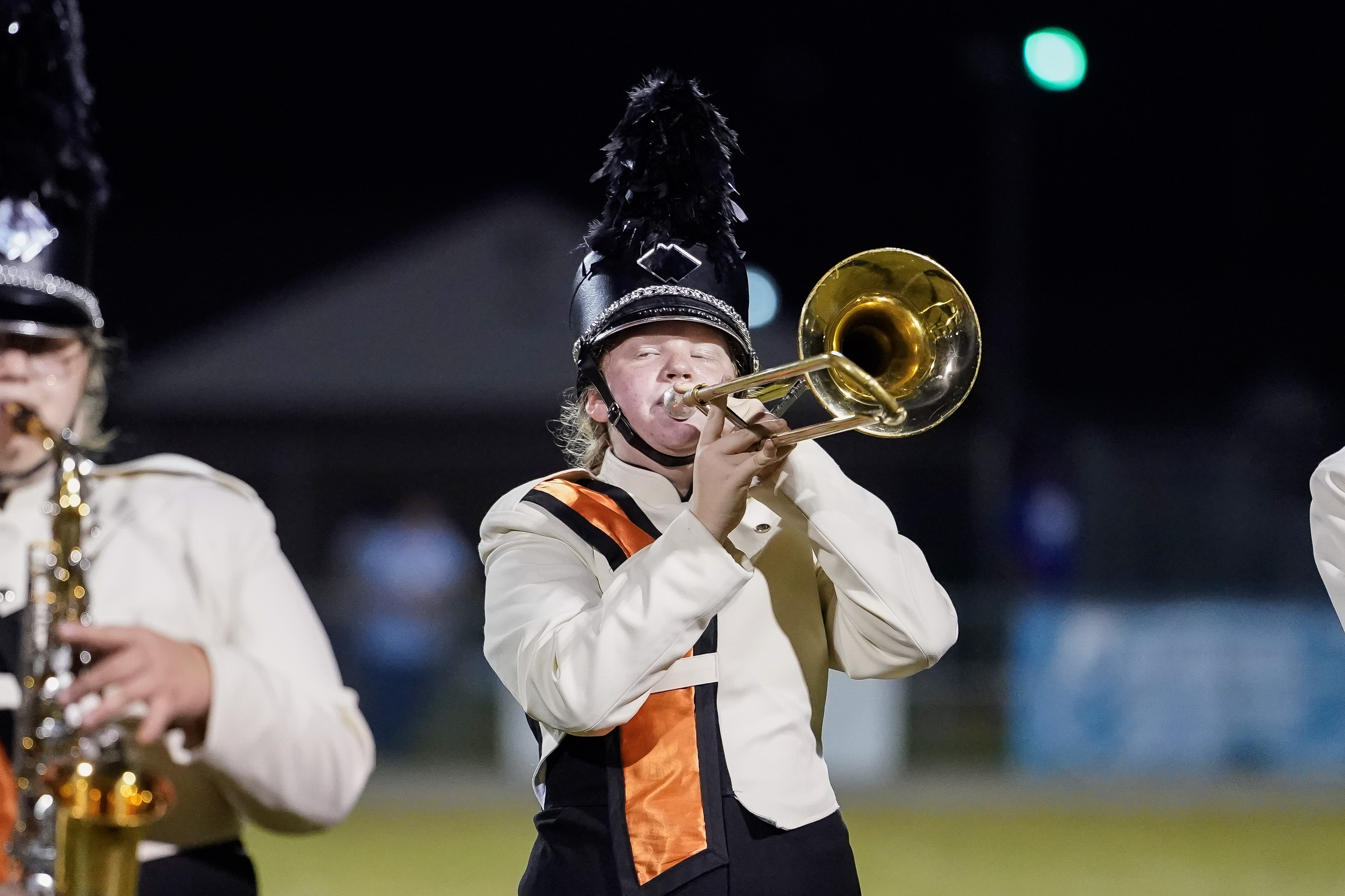 A marching band member in a white and black uniform with an orange stripe plays a trombone at night. The person wears a tall, feathered hat, and other band members are partially visible in the background.
