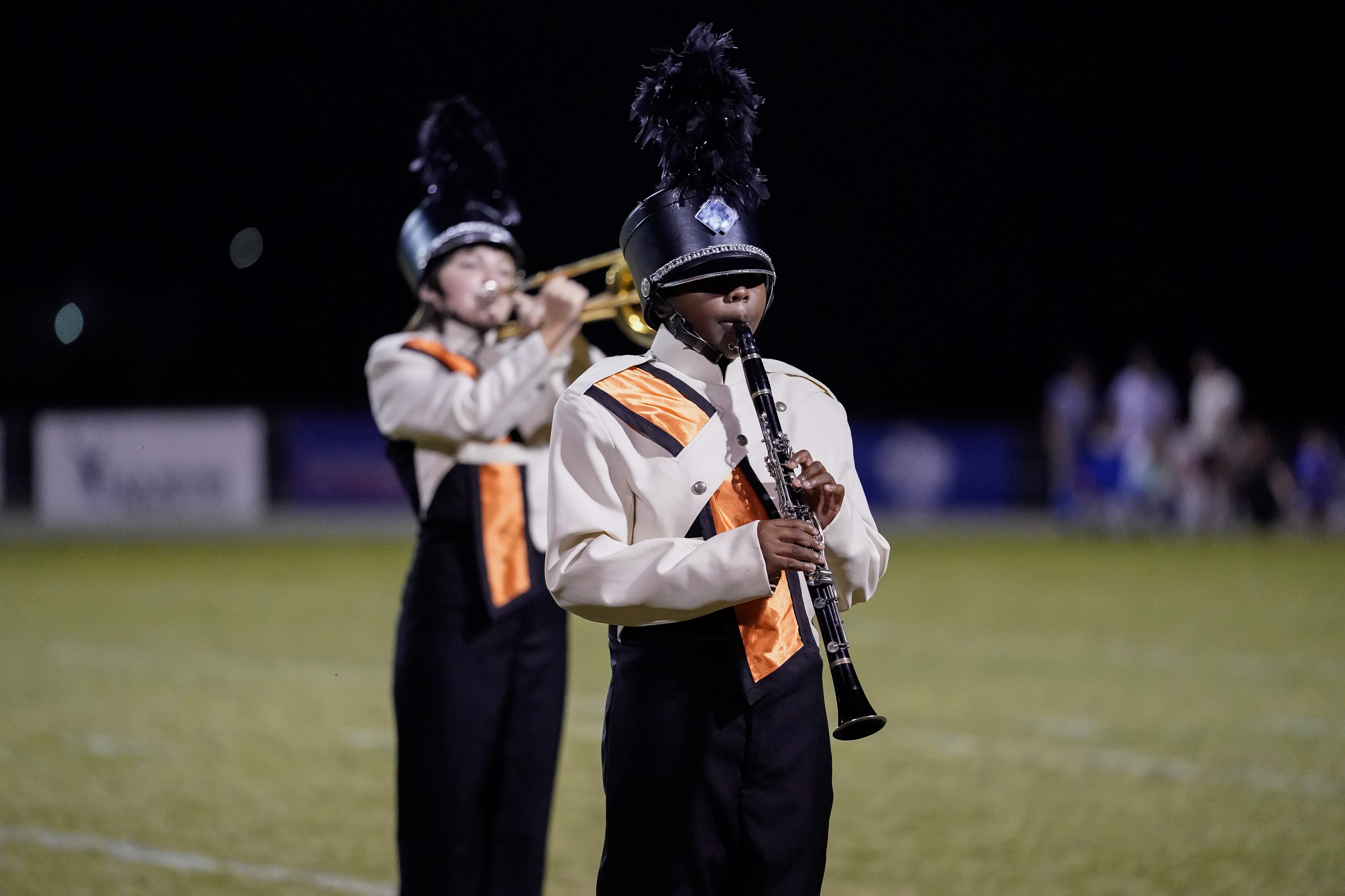 Two marching band members perform on a field at night; one plays the clarinet in the foreground, wearing a tall feathered shako, and the other plays a brass instrument in the background. Both wear matching uniforms.