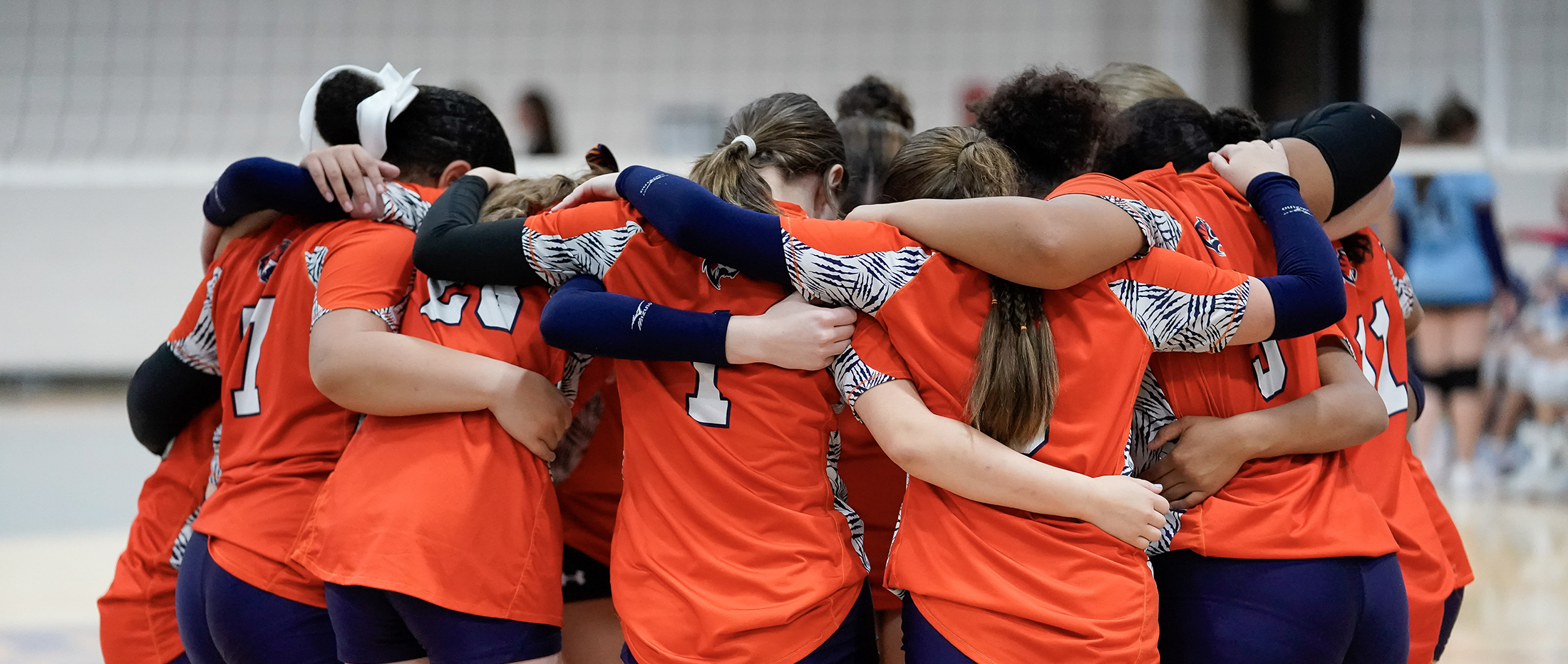 Middle school volleyball players wearing orange and blue uniforms huddle with heads bowed and arms wrapped around each other