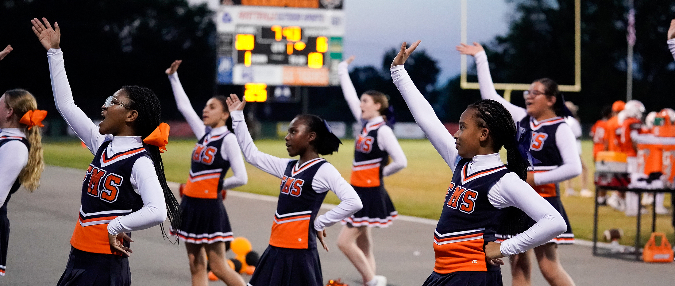 middle school cheerleaders in blue and orange uniforms hold up their arms during a cheer