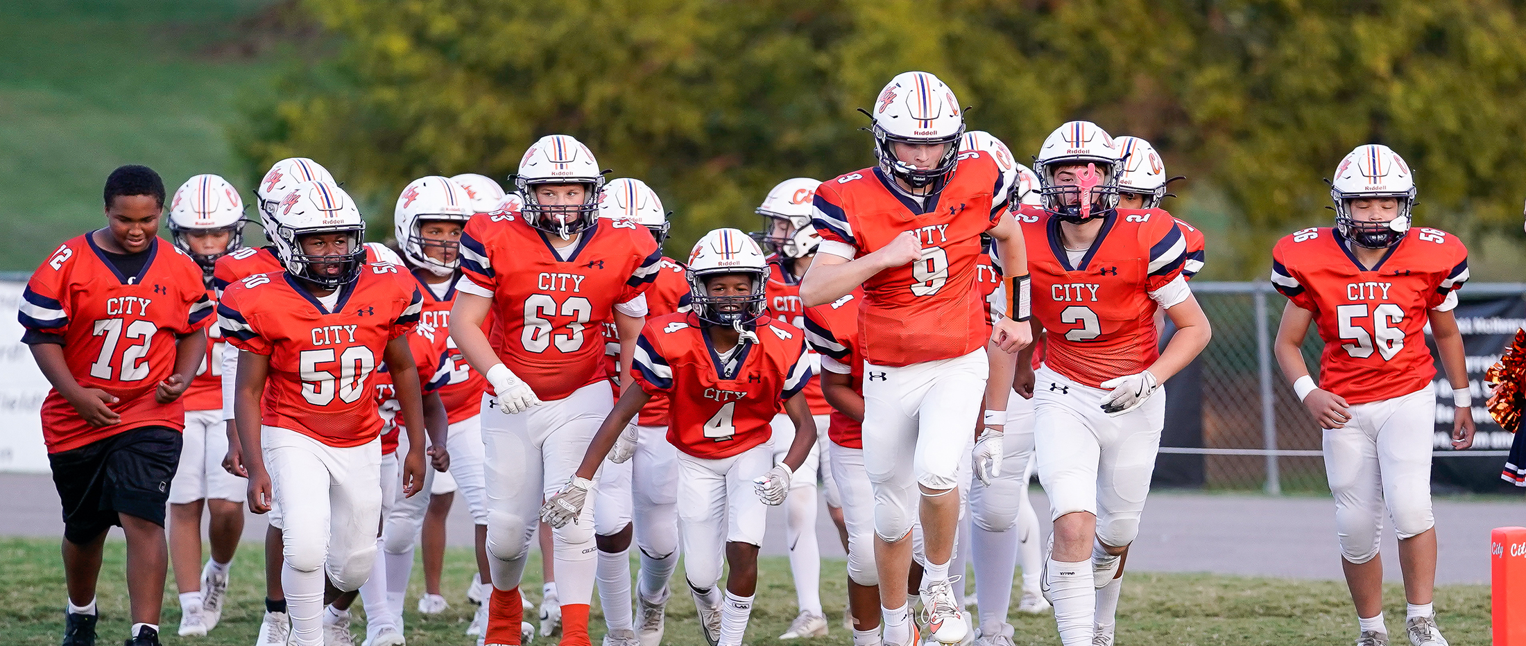 middle school football team wearing orange jerseys, white pants and white helmets run onto the field before a game