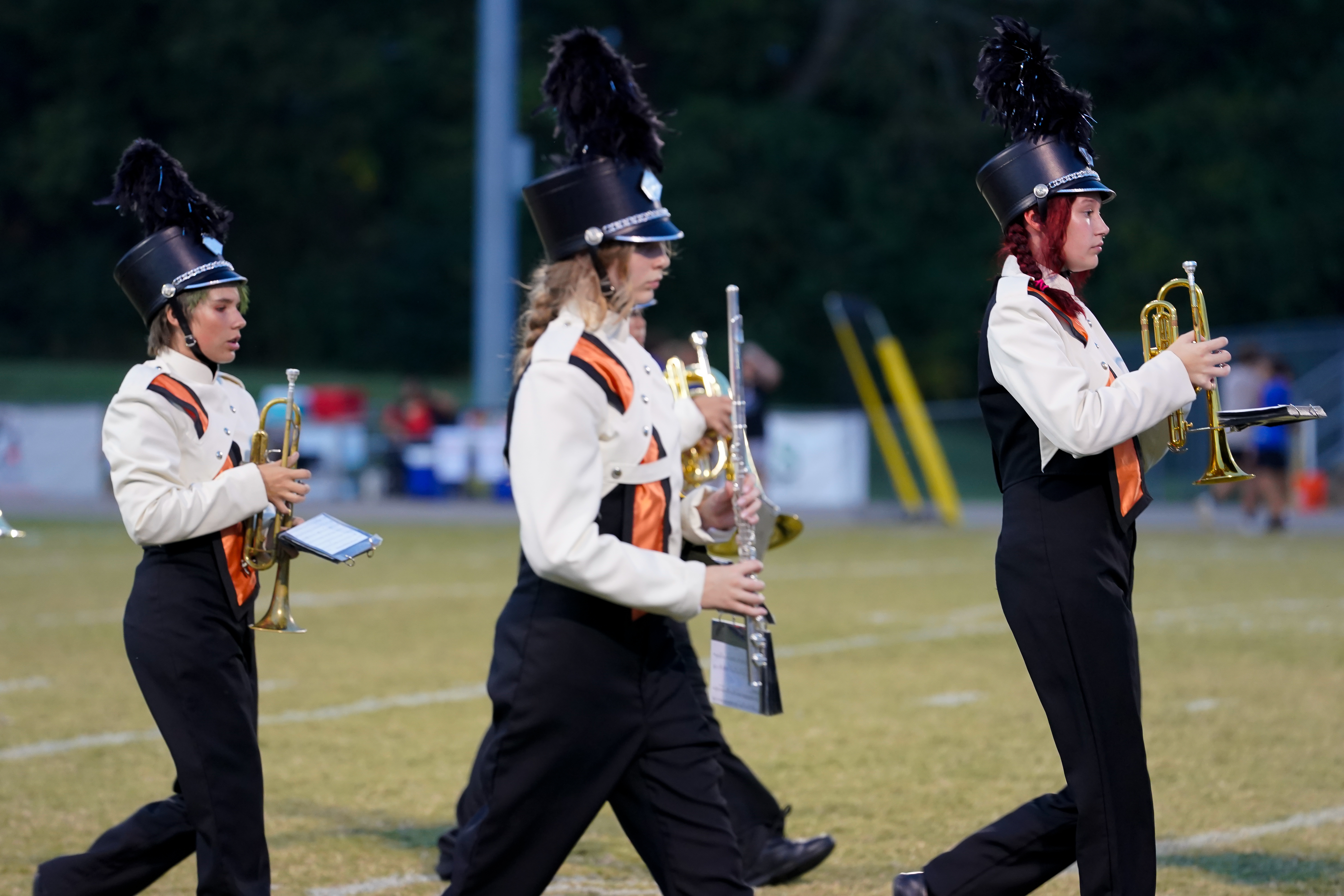 Three Fayetteville High School marching band members in white and black uniforms walk across the field, holding trumpets and a flute with feathered shakos.