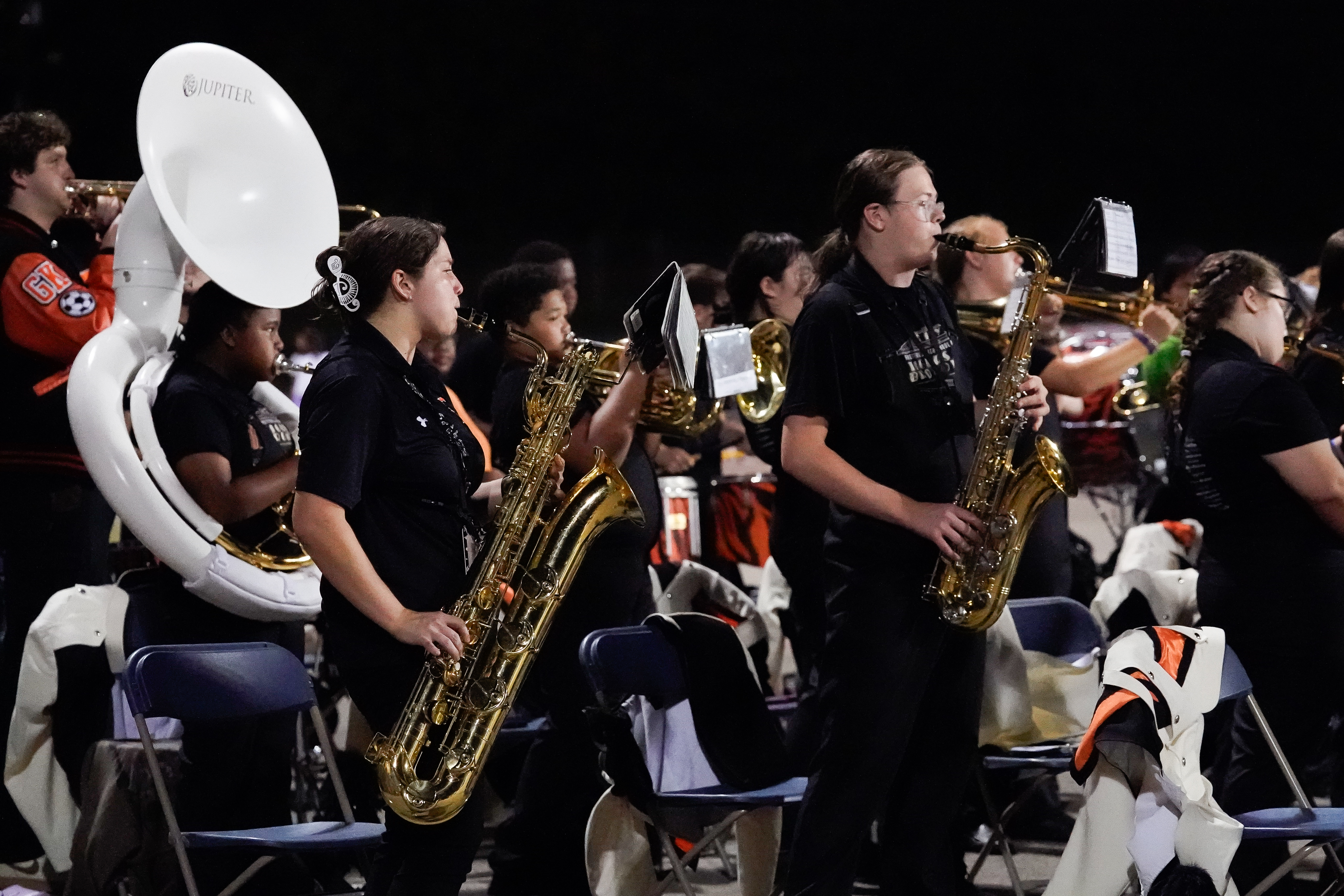 A group of band members in dark uniforms play brass instruments, including saxophones and a sousaphone, during a performance at an indoor event.