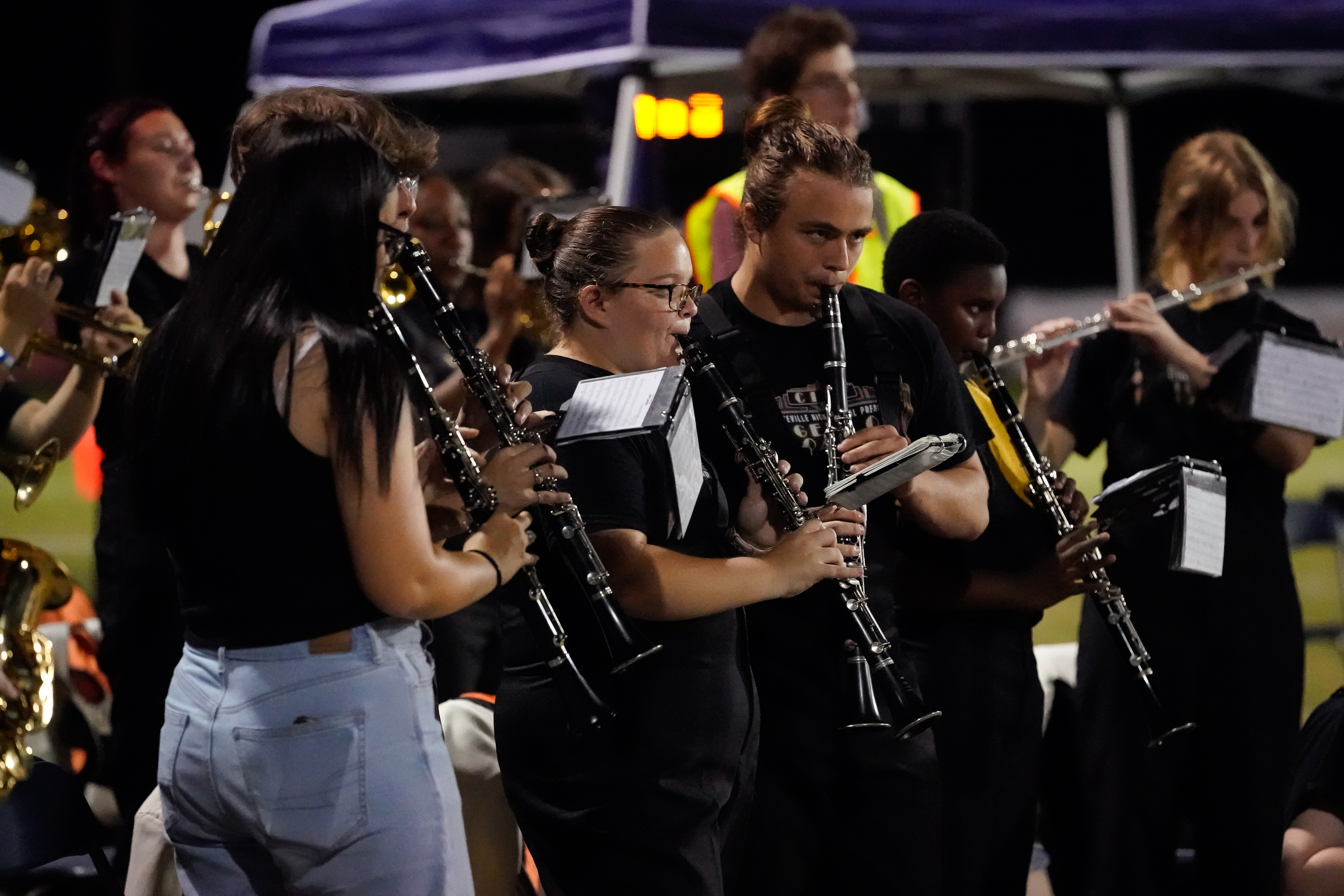 A group of young musicians in black shirts play clarinets and flutes during a nighttime outdoor performance, with music stands in front of them and a tent in the background.