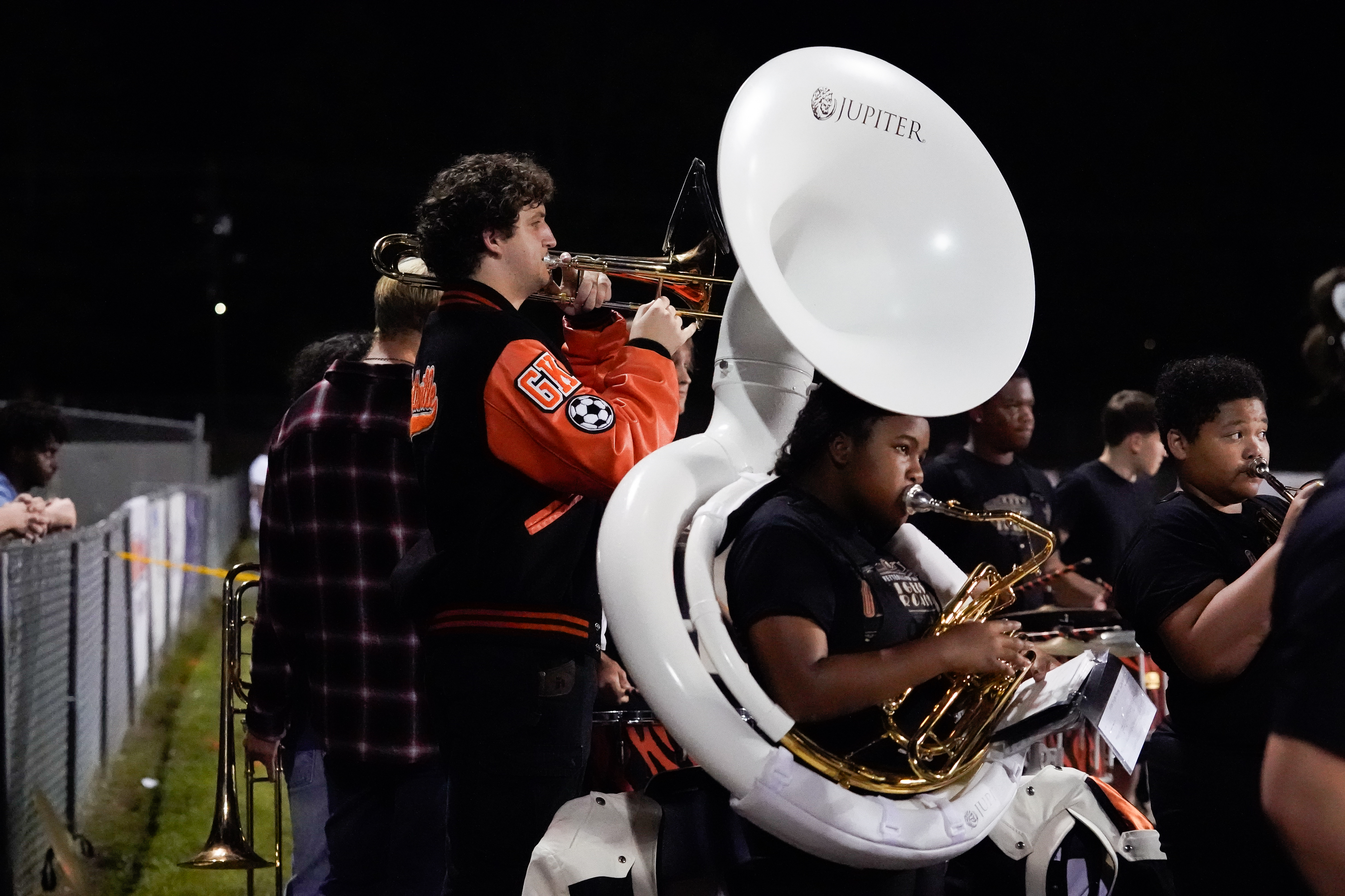 A high school marching band performs at night. One student plays a sousaphone while wearing a black shirt, and another in a letterman jacket plays a trombone. Other band members are visible in the background.