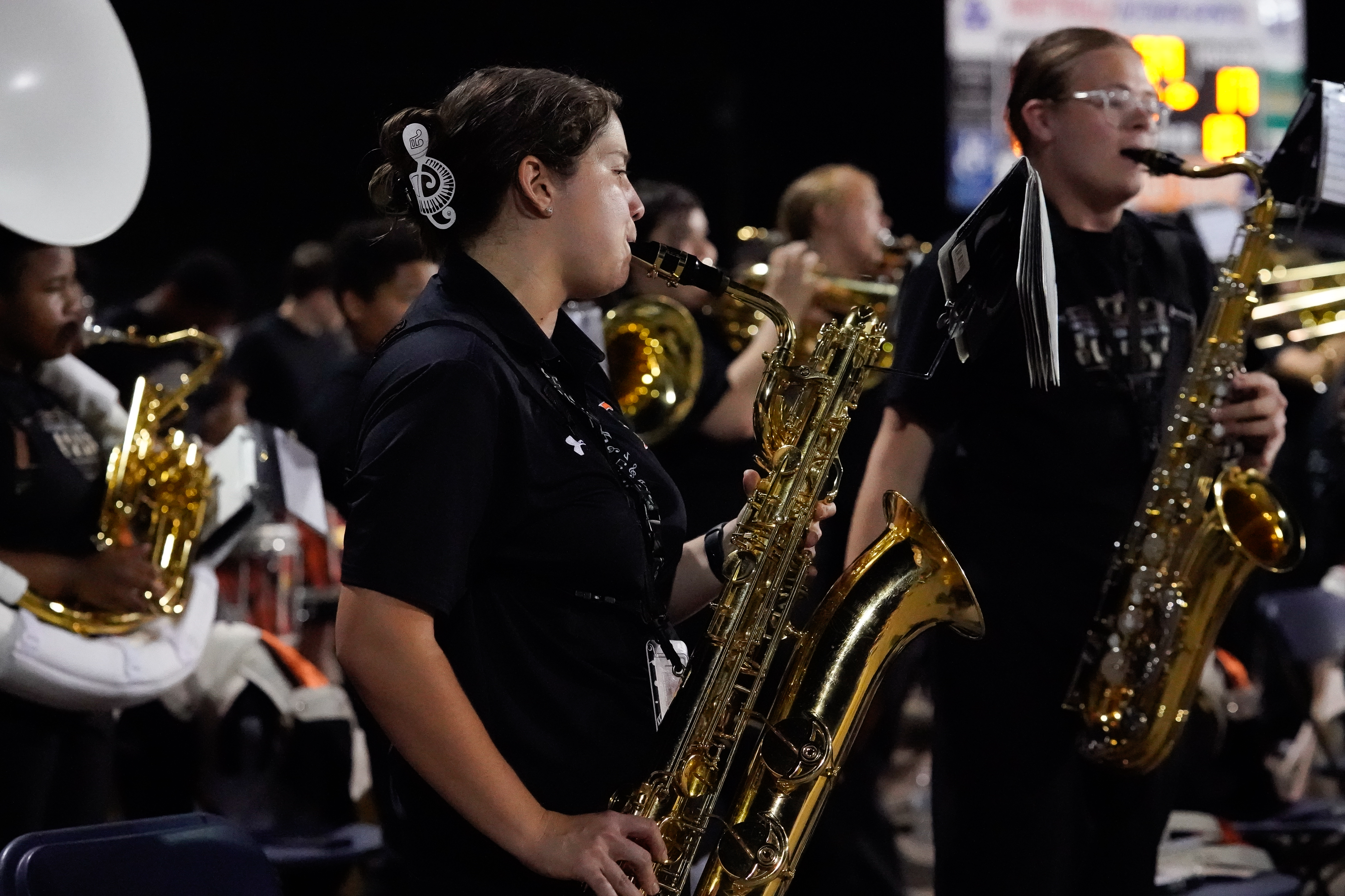 A school marching band performs at night, with students standing and playing brass instruments, including saxophones and a sousaphone, while wearing matching uniforms. Empty folding chairs and instrument cases are visible.
