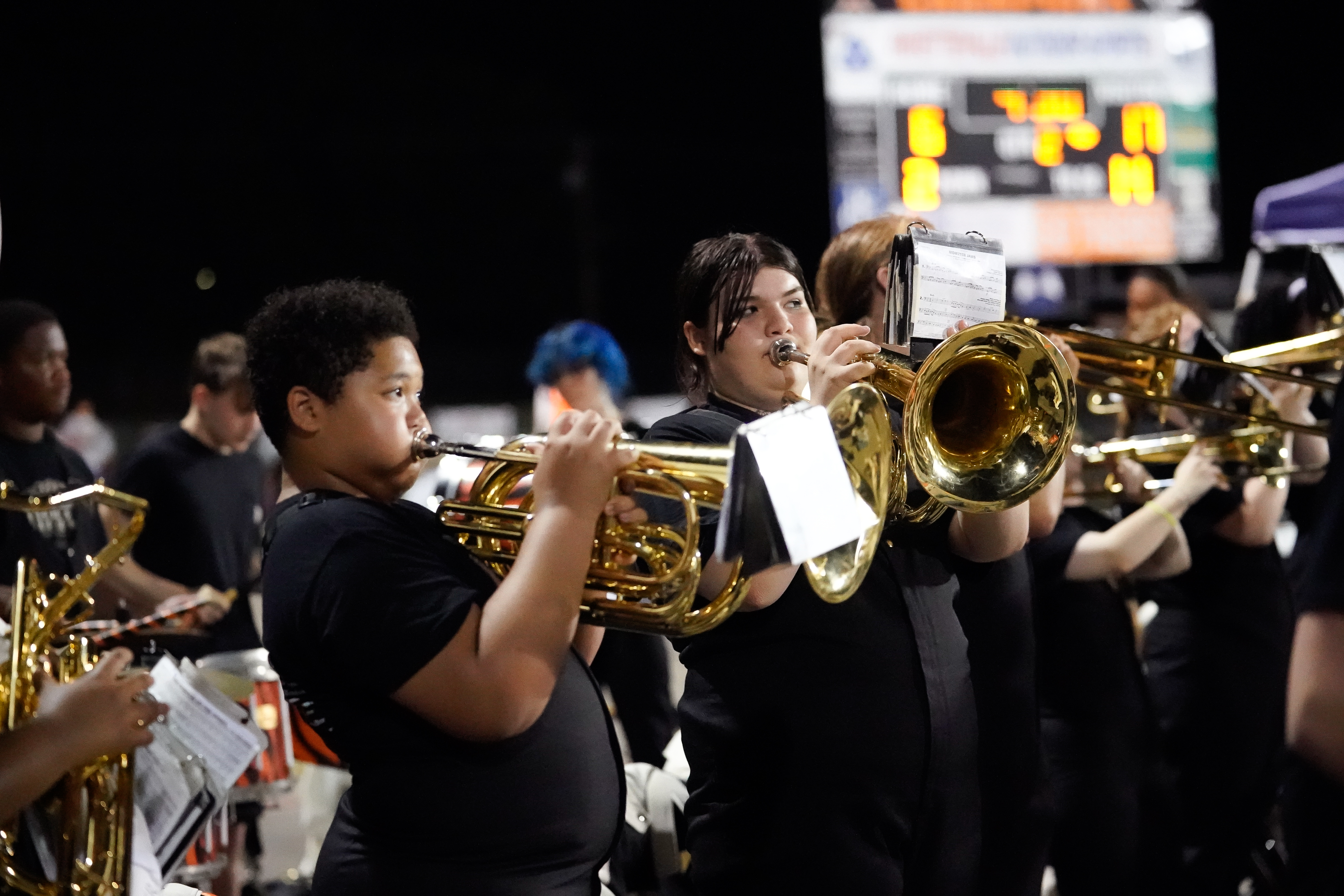 A group of marching band students in black uniforms play brass instruments at night, with a scoreboard visible in the blurred background.