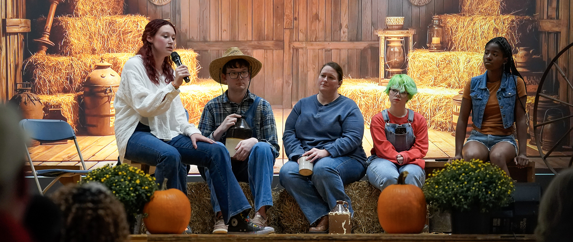 a girl seated on a haybale singing with others watching dressed in Hee Haw costumes